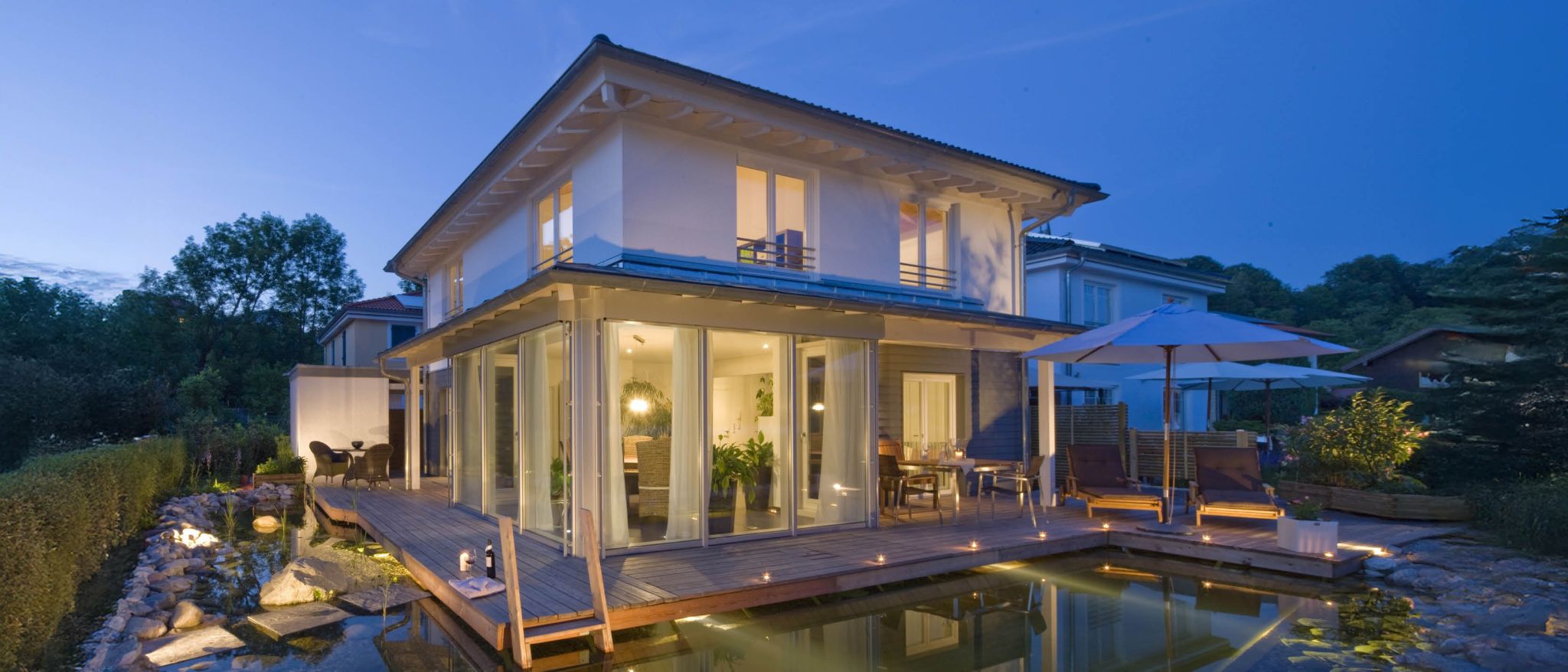 Modernes Haus mit Glasanbau, Holzdeck, Teich und Beleuchtung am Abend mit blauem Himmel.