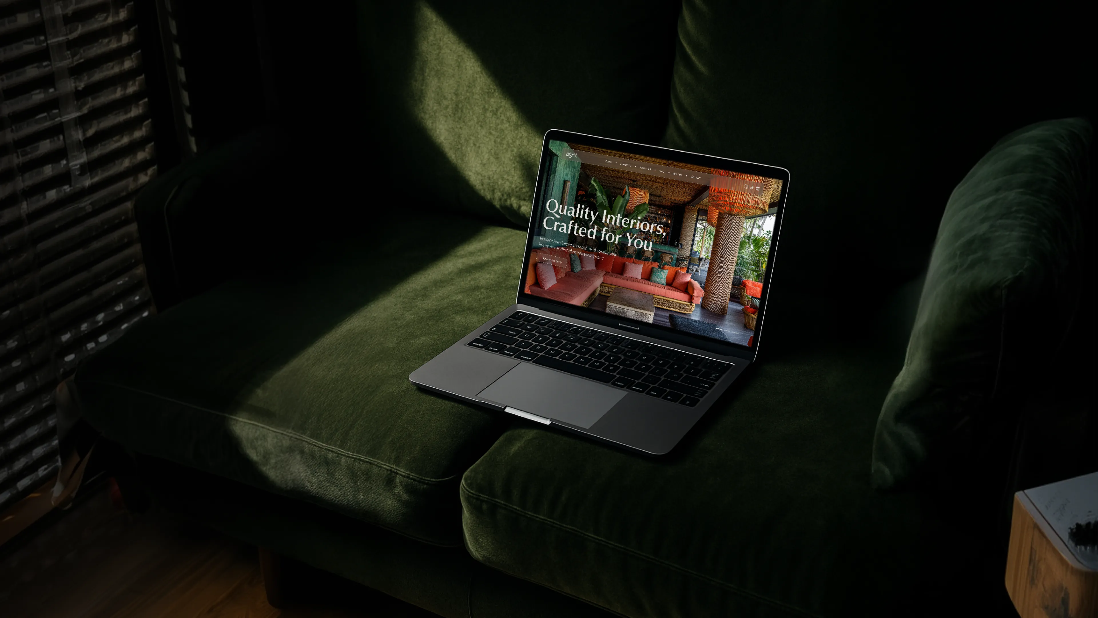 MacBook laptop open on a dark green velvet armchair with dappled window light, displaying the Objet website hero with the headline "Quality Interiors, Crafted for You" over a richly colored interior photograph with terracotta sofas and tropical plants