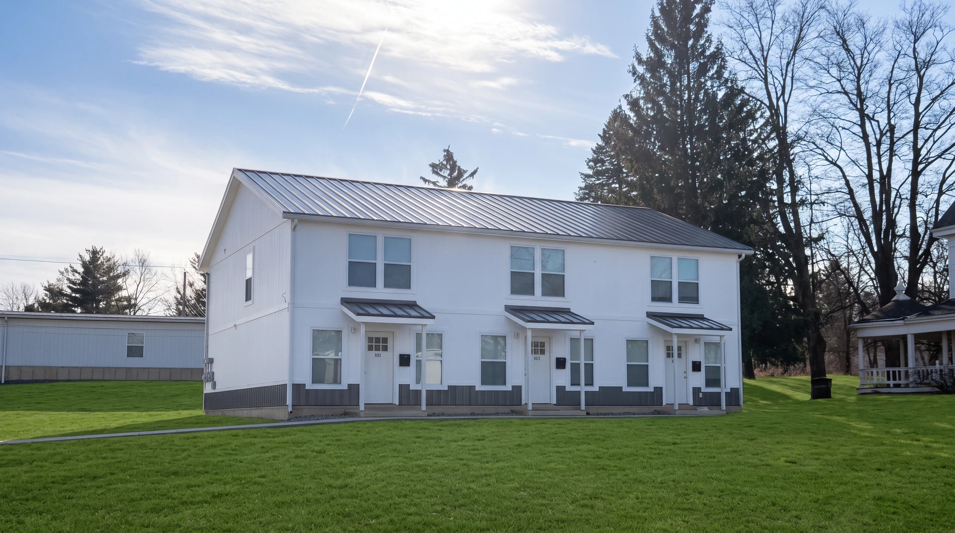 A white three-unit triplex with a standing-seam metal roof and three separate covered entries, set back on a wide lawn