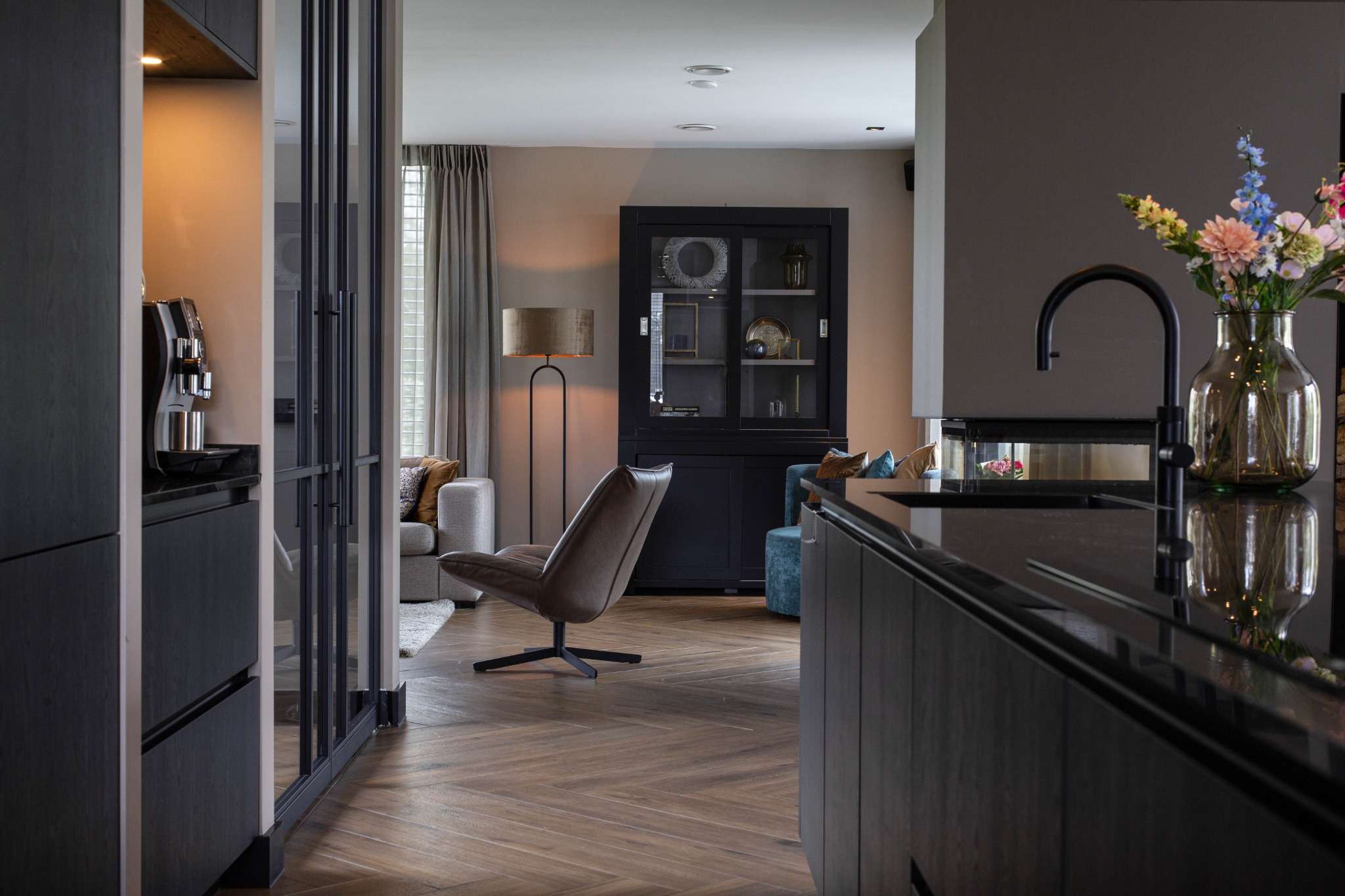 Doorkijk van keuken naar woonkamer met loungefauteuil, donkere kast en warme sfeerverlichting.