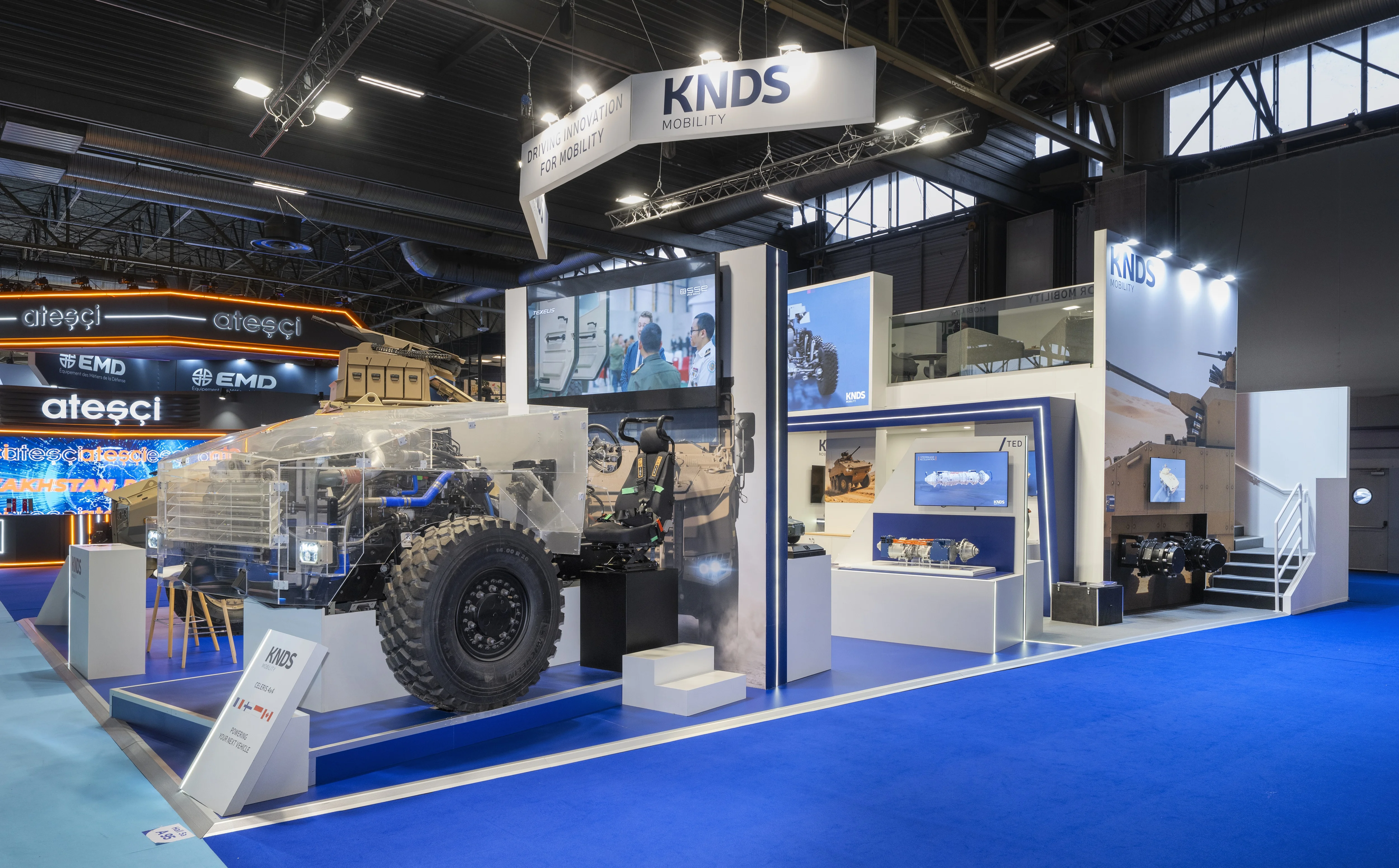 Stand KNDS Mobility conçu et réalisé par Expace à Eurosatory 2026 – espace sur-mesure défense avec blindé SCATA, châssis Celeris écorché, bandeau suspendu « Driving Innovation for Mobility » et lounge à l'étage.
