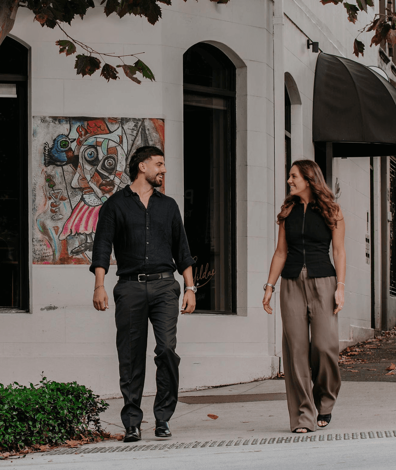 Two smiling people walk together on a city sidewalk.