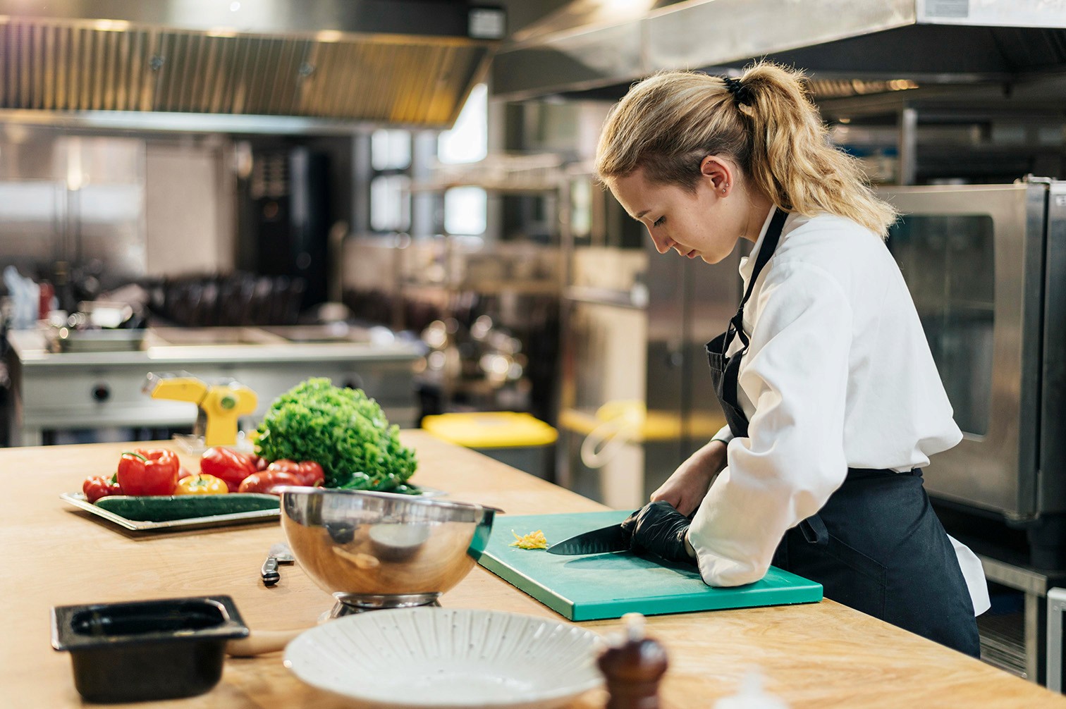 A female chef in a white uniform and black apron, slicing a vegetable on a wooden counter with various fresh ingredients.