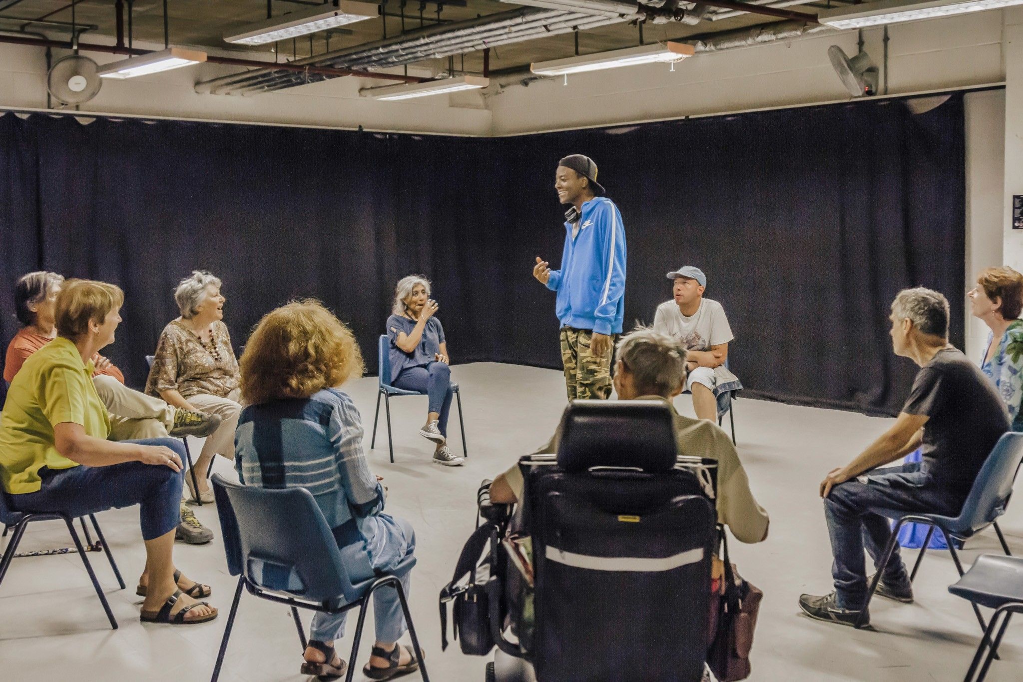 Young man smiling in drama studio, with a mixed-age audience seated in a circle around him