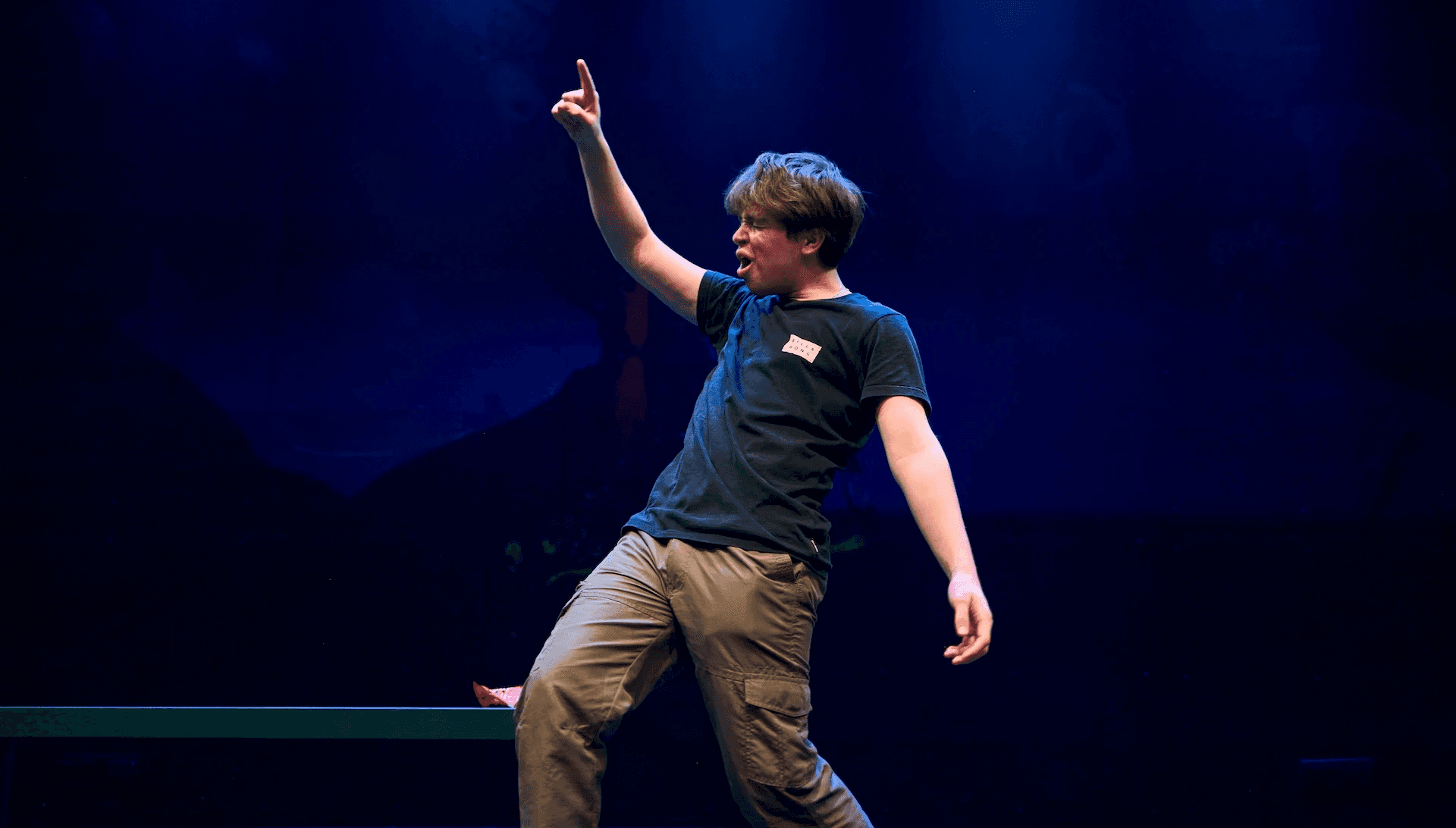 A young performer stands centre-stage under blue lighting, striking an exaggerated pose with one arm pointed upward and their mouth open mid-expression. They wear a black T-shirt and cargo pants, with a prop or small item resting on a bench beside them.