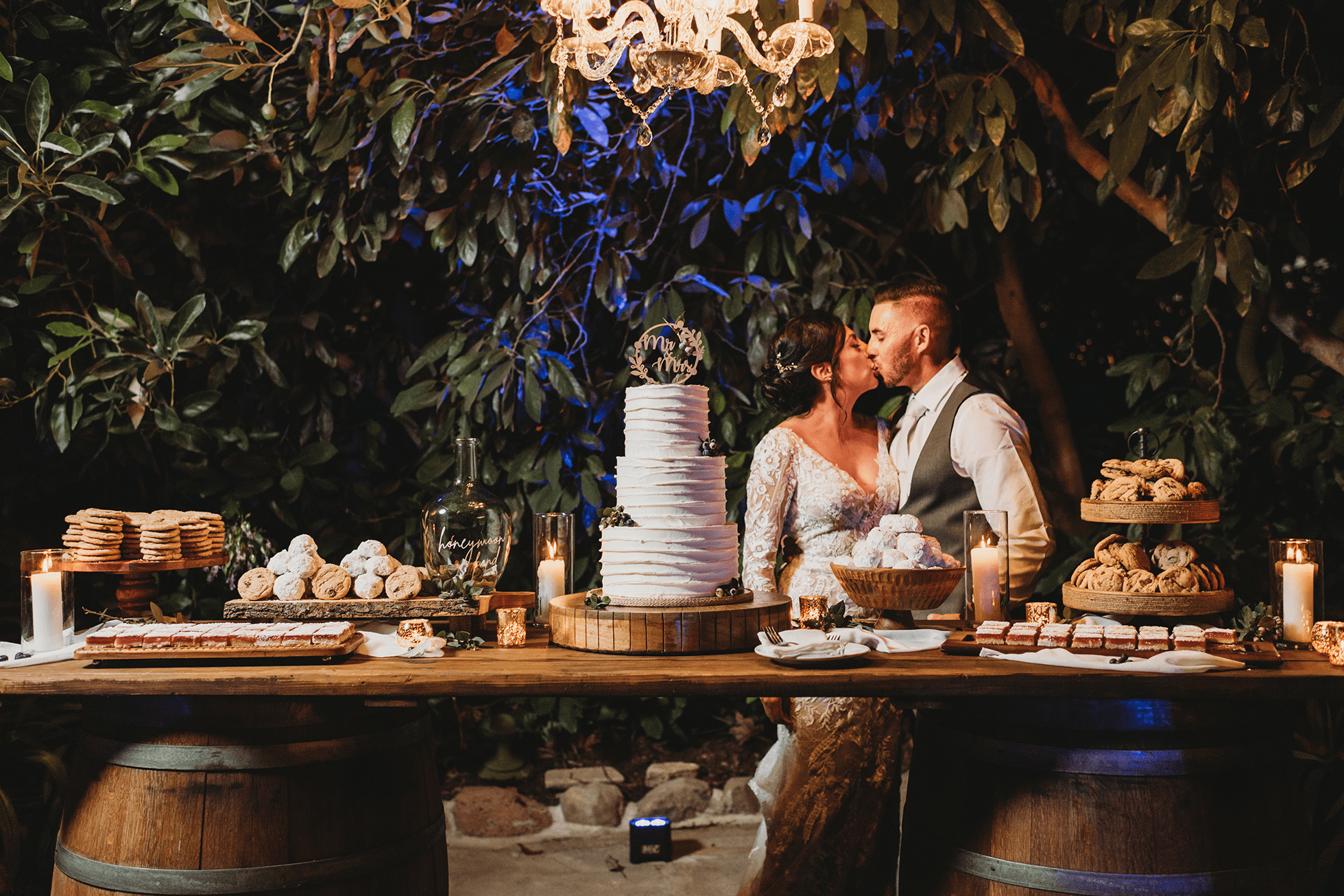Bride and groom kissing behind dessert table at night with blue uplighting