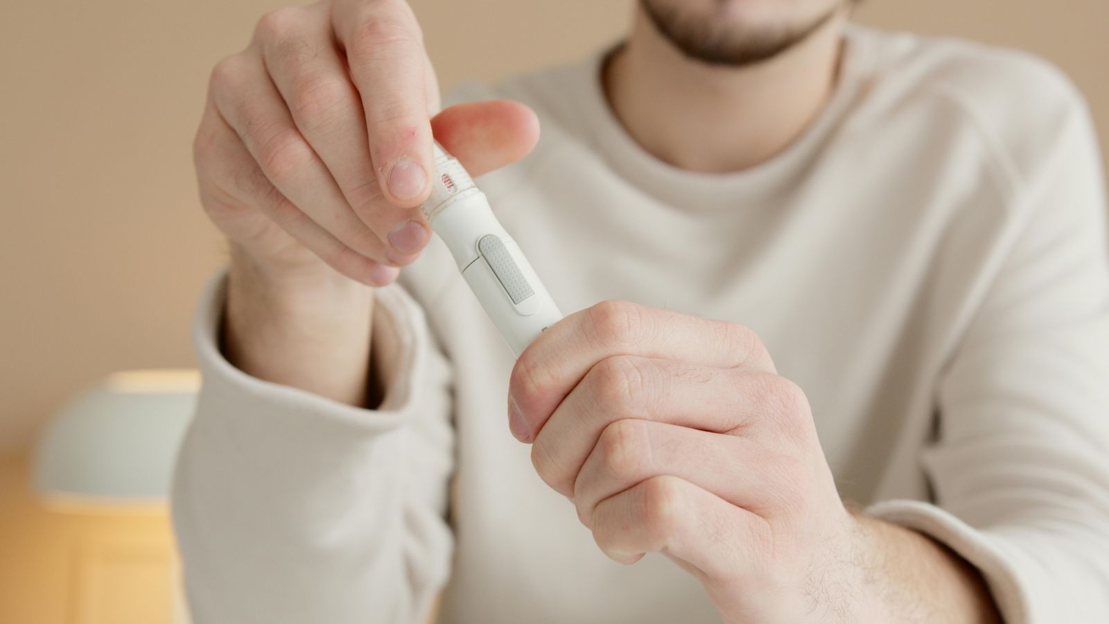 A person holding a white lancing device for blood glucose testing, with a textured grip, button mechanism, and adjustable depth dial, preparing to use it for obtaining a small blood sample.