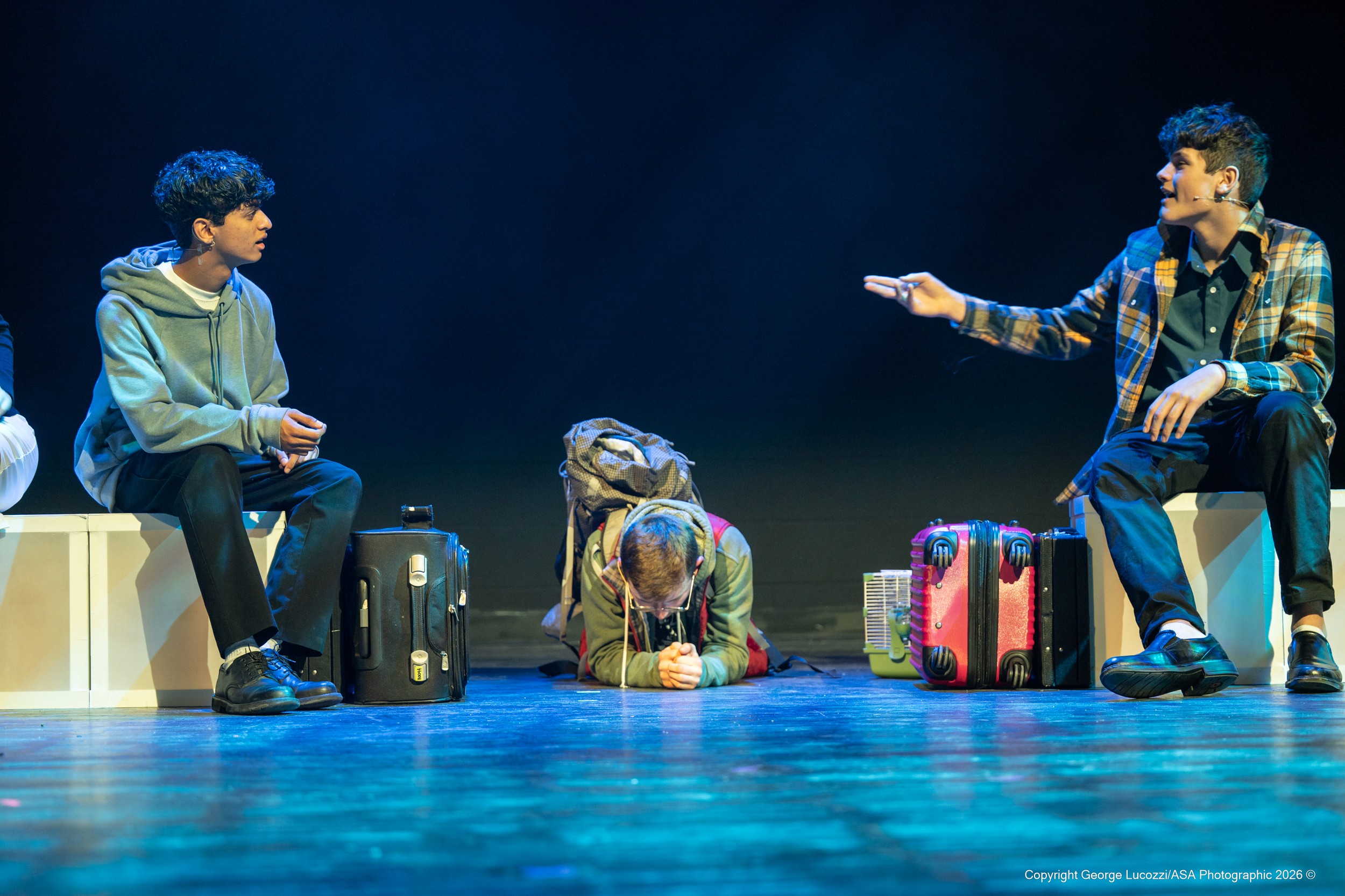 Christopher crouches center frame among blocks and suitcases, flanked by two “Voices” portraying cheerful, intoxicated passengers; cool blue and soft warm light blend to create a lively yet isolating transit atmosphere.