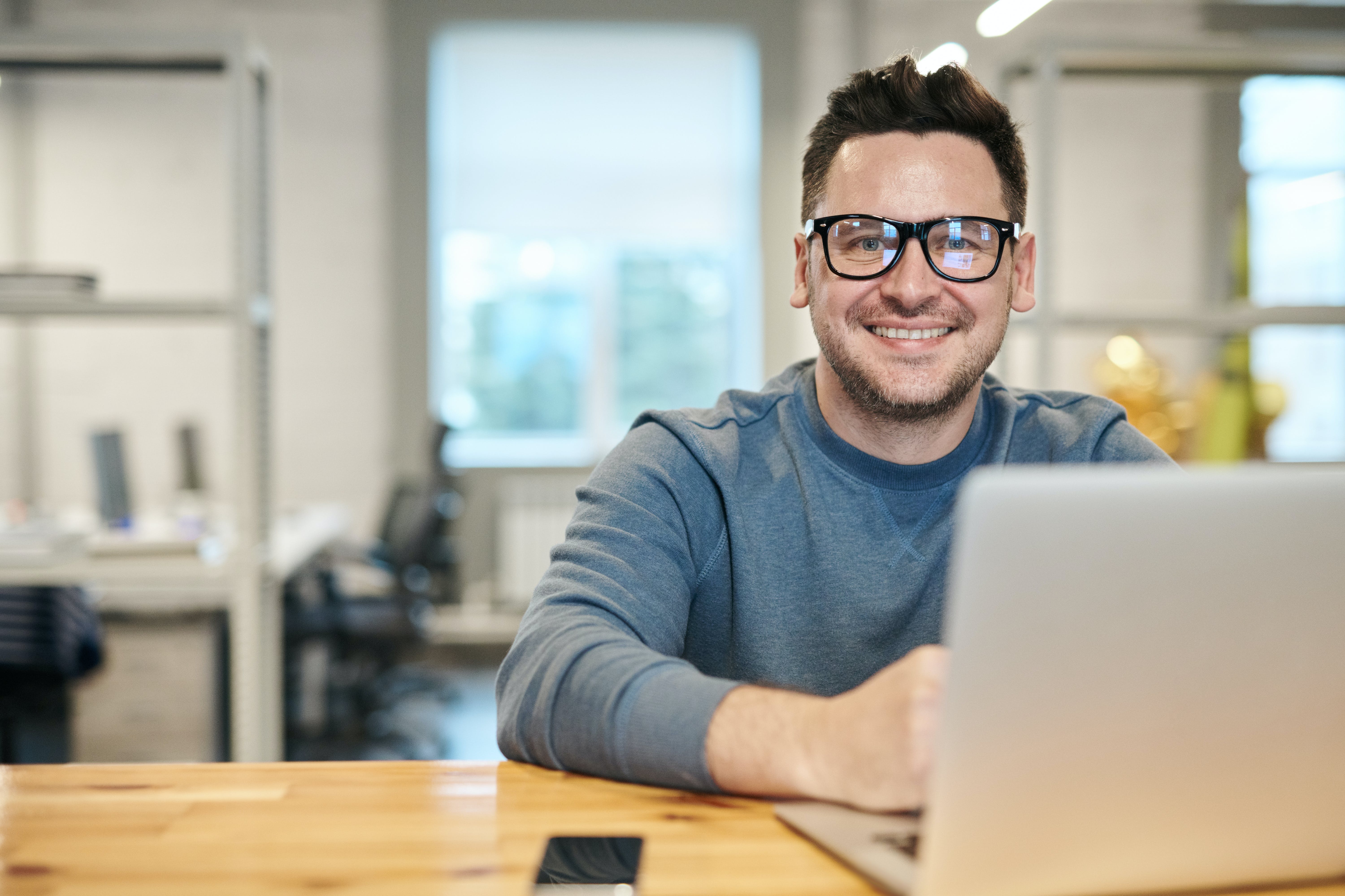 A smiling man wearing glasses sitting at a wooden table with a laptop, looking at the camera.