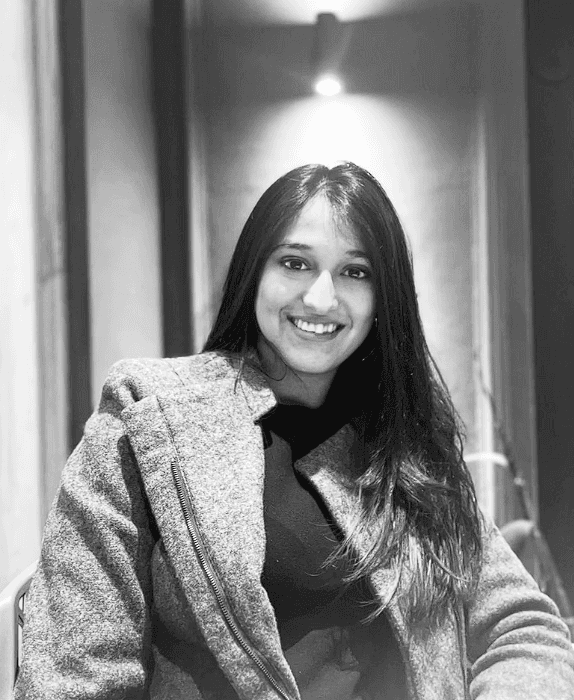 A smiling person with curly hair sits at a desk, looking at a laptop against a dark background.