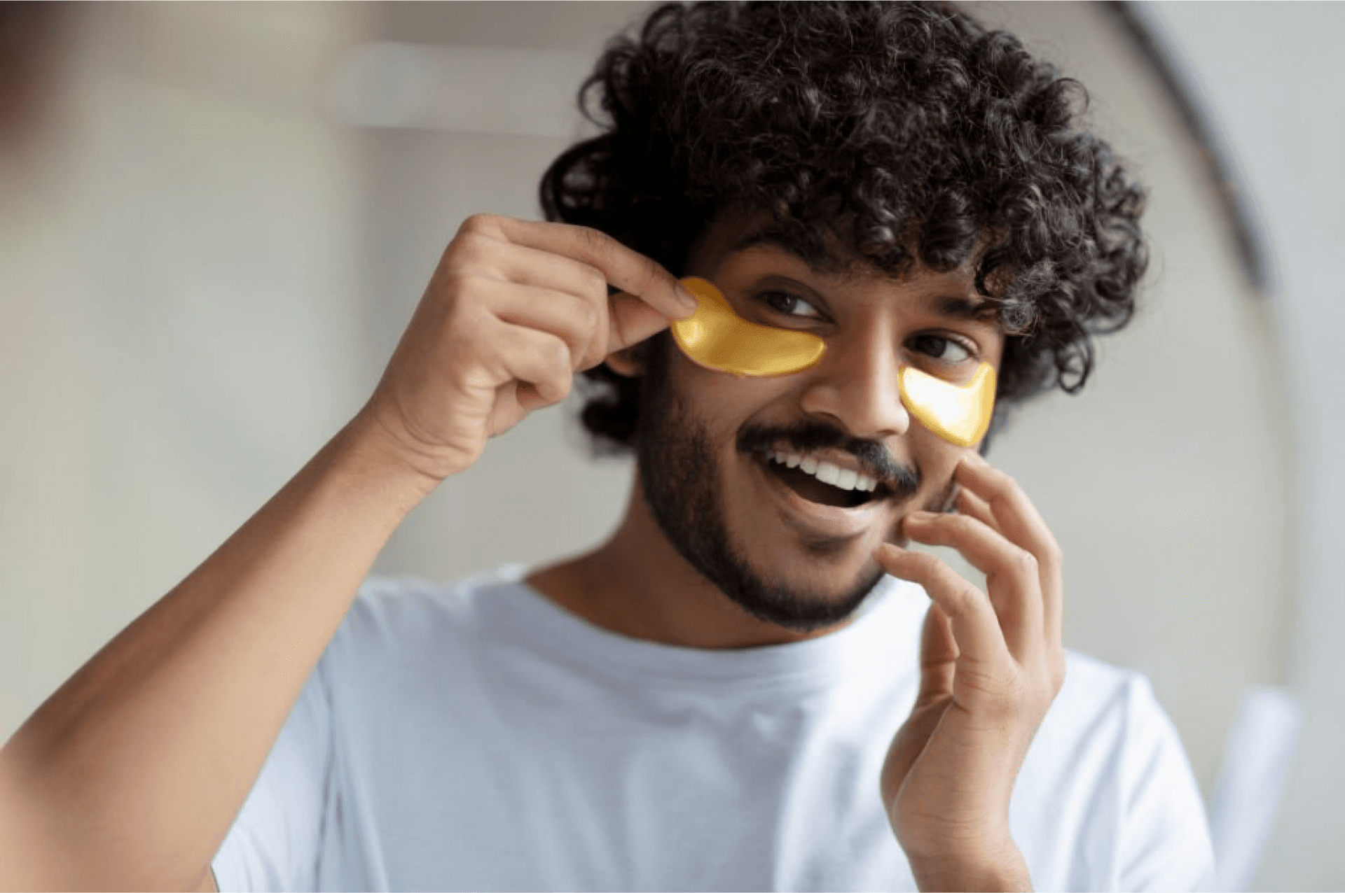 Man smiling while applying golden under-eye patches for skincare.