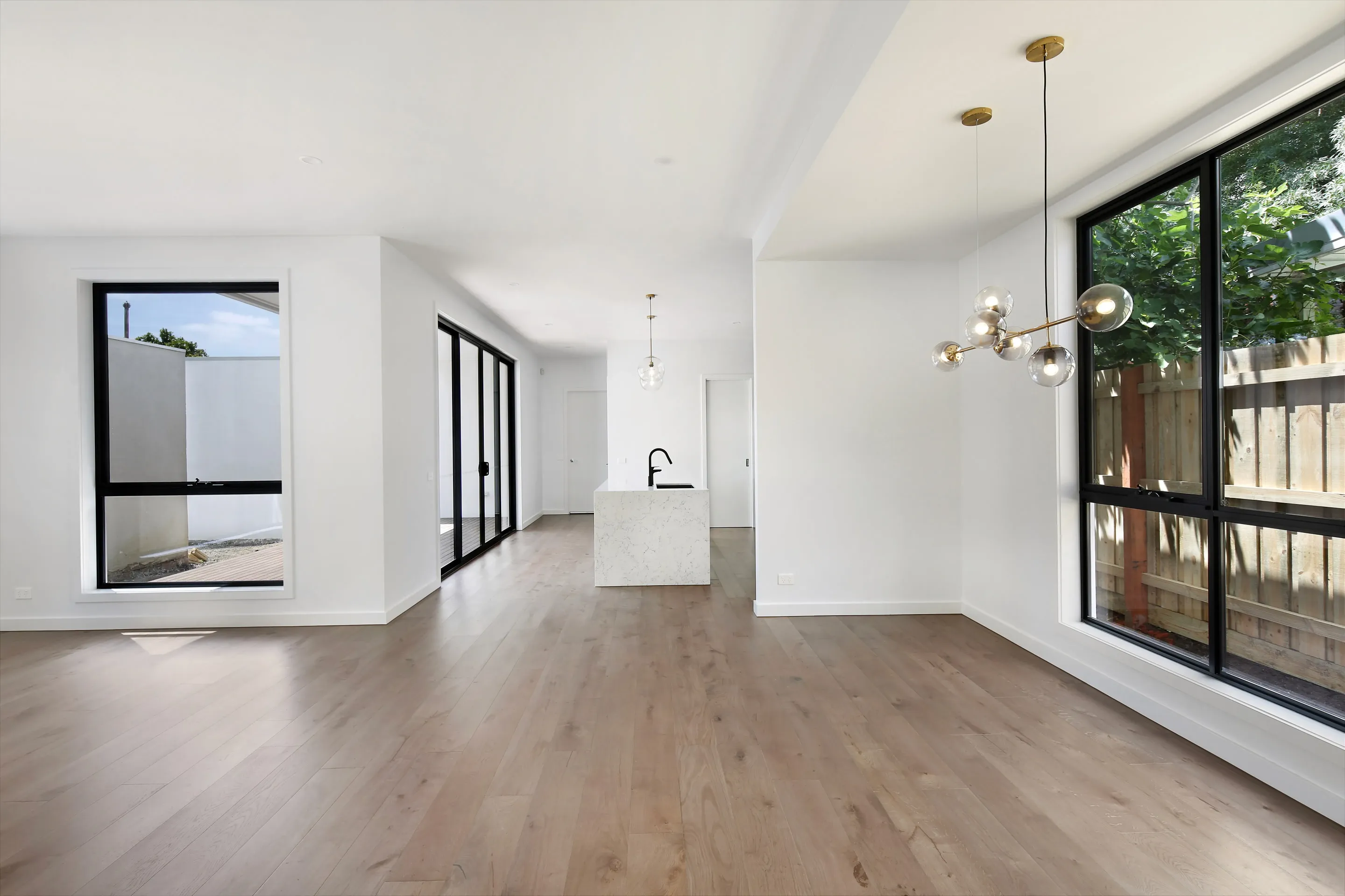 Open living and dining with white walls, oak floors, a brass-and-glass globe chandelier, black-framed windows to a fence and a light court, and a stone island with black tap beyond