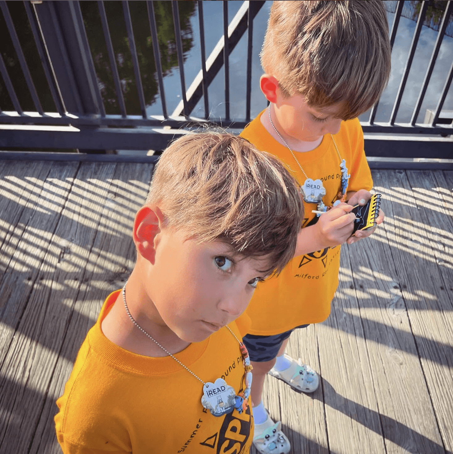 Two young boys in yellow shirts stand on a wooden deck, looking down at their phones. Bright sunlight illuminates the scene.