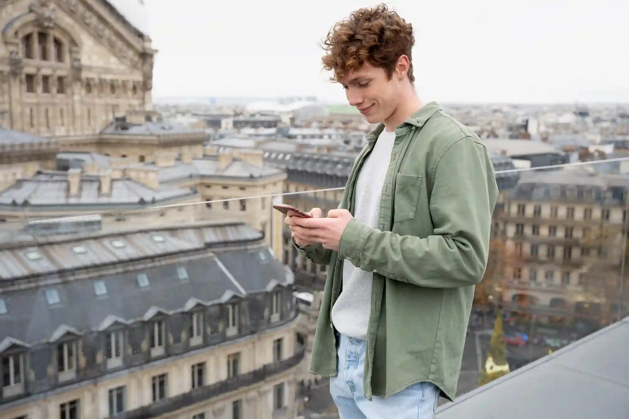 A young man smiles while using his smartphone on a rooftop overlooking the historic city skyline of Paris.