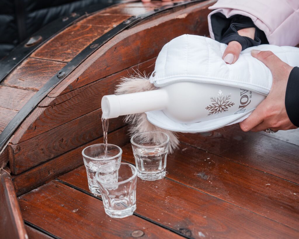 Snow Tequila being poured from a bottle in a white padded cover into small glasses on a wooden table