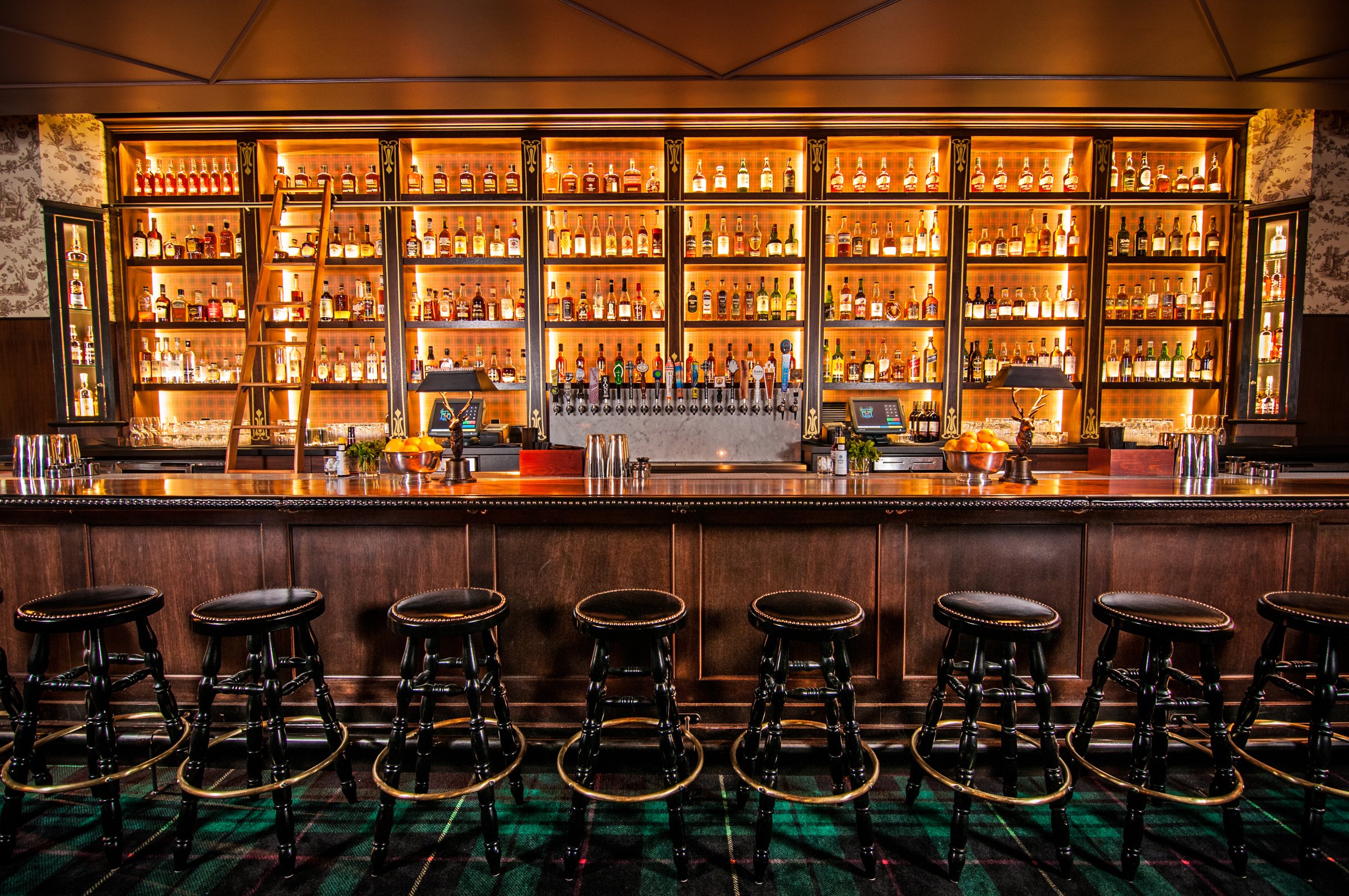 Long dark timber bar with black stools and illuminated bottle shelves above a tartan-patterned floor.