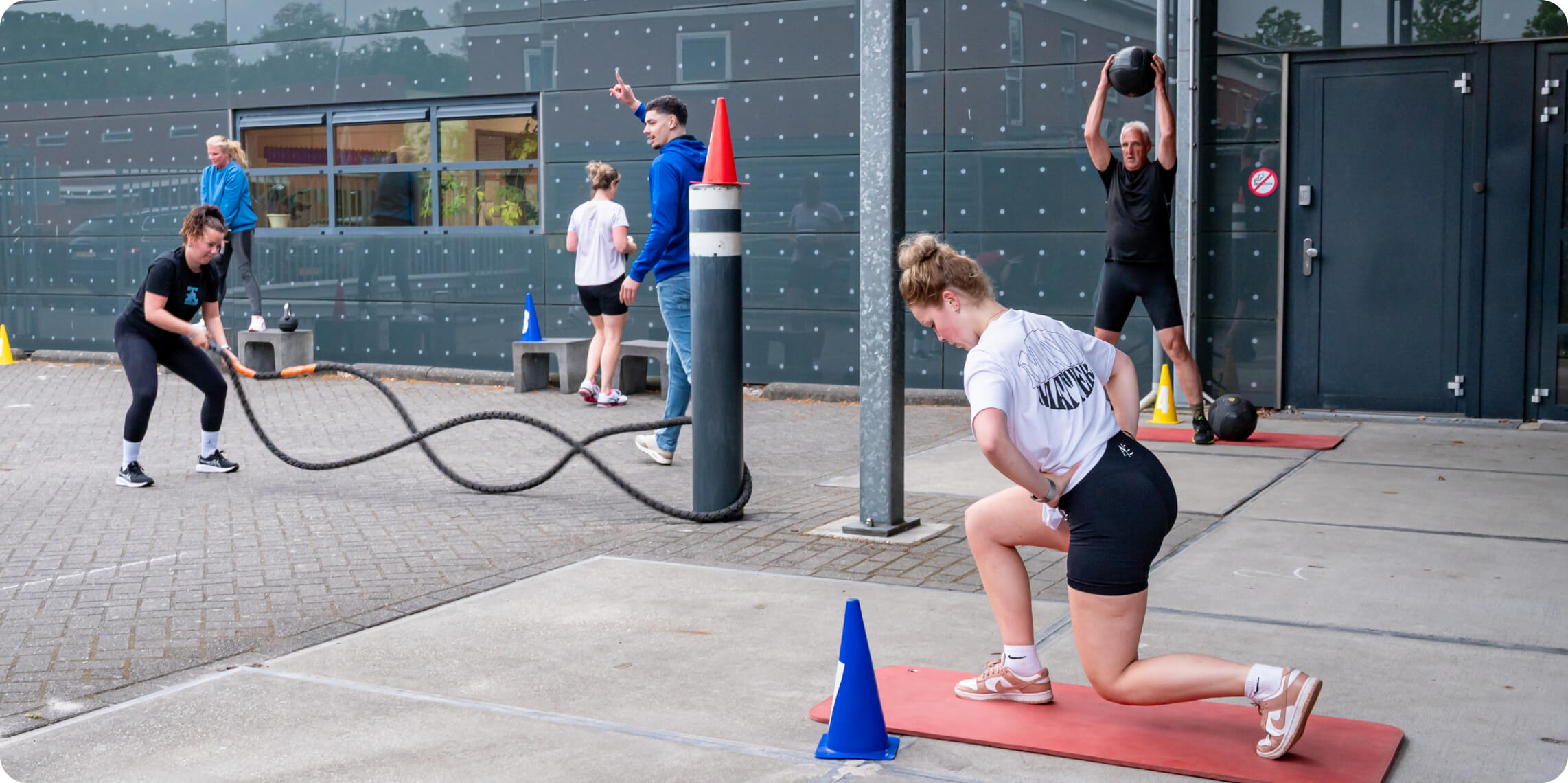 Outdoor fitness bootcamp with people using battle ropes, performing lunges, and lifting medicine balls near a modern building with reflective glass.