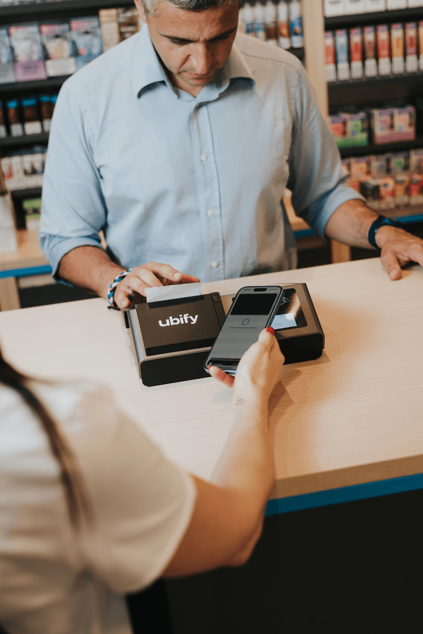 Cliente effettua un pagamento digitale con smartphone sulla Cassa Ubify, assistito da un commerciante in negozio.