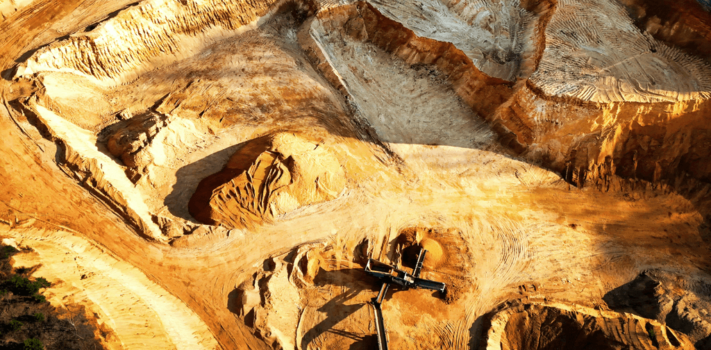 An aerial, high-angle view of an active open-pit mine or quarry during late afternoon, bathed in warm, golden sunlight. The landscape is deeply excavated, revealing carved ridges, terraces, and steep slopes of earth in shades of ochre, tan, and reddish-brown. Tire tracks mark the dusty ground below. In the lower-center section of the pit, heavy industrial mining equipment, including a conveyor belt system, sits near small piles of excavated material. Deep shadows stretch across the textured, uneven terrain, emphasizing the massive scale of the excavation.