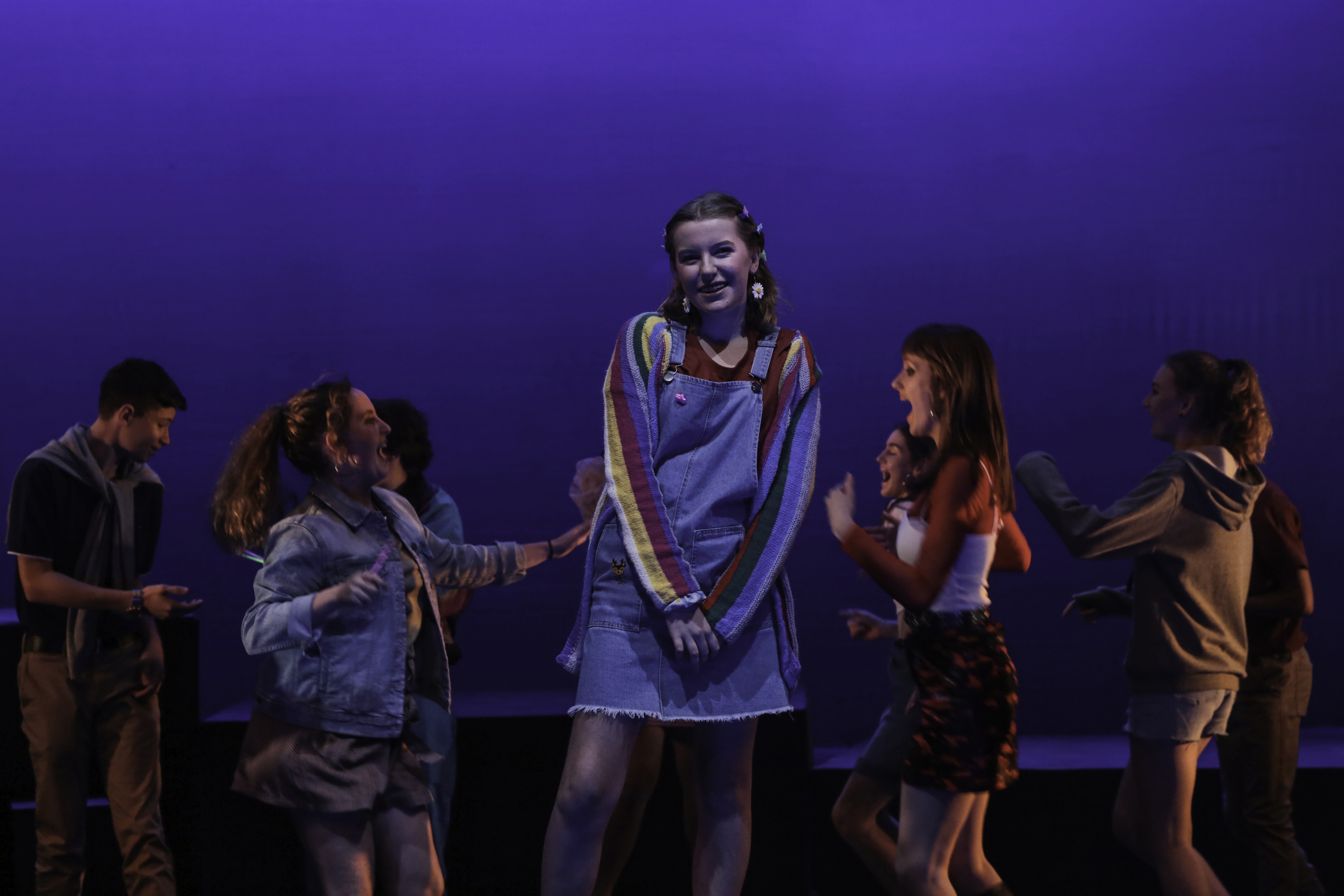 A teenage girl in denim overalls and a rainbow cardigan stands shyly at a party while other teens dance around her. Blue lighting and lantern props create a dreamy nighttime setting.