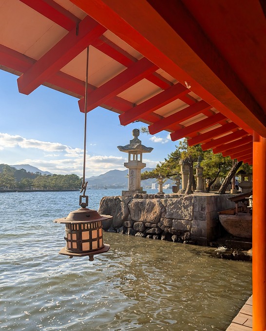 The view from inside a Japanese temple, with the roof falling across the image. A lantern hangs in front of sculptures and in the distance, mountains.