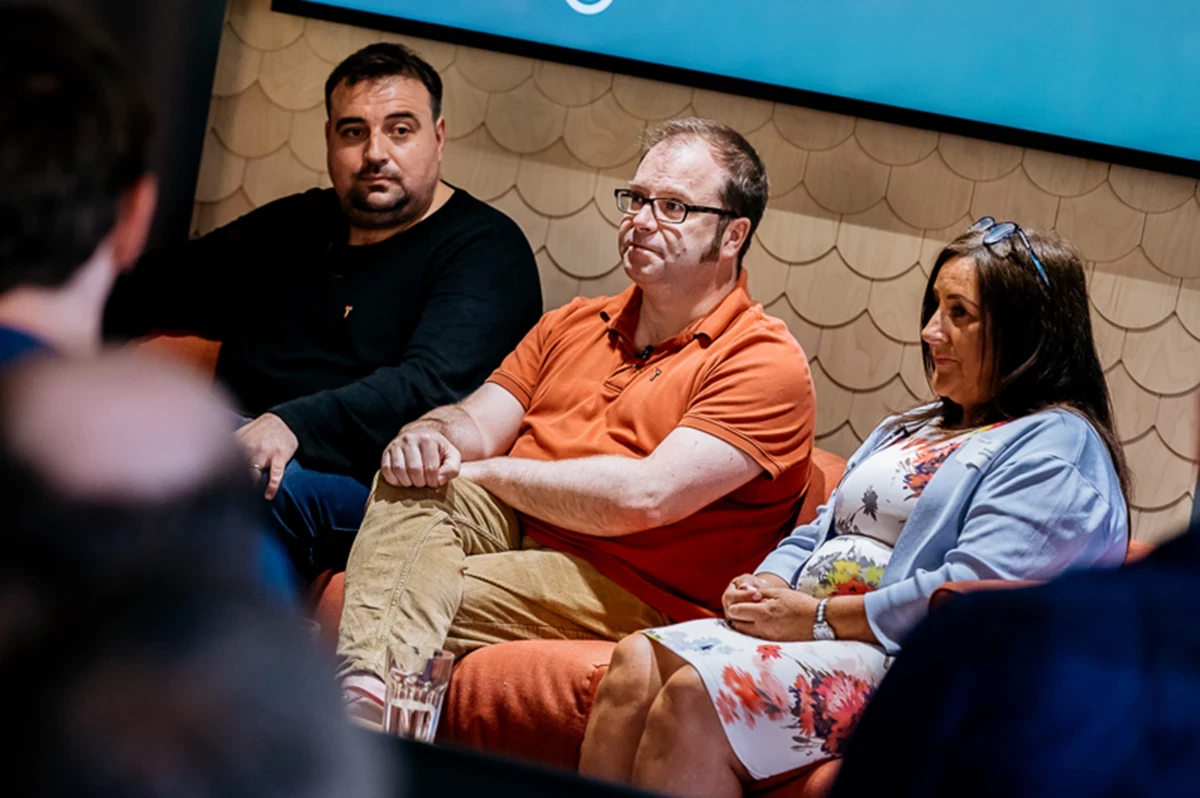 Three people seated on a sofa during a discussion or panel event, with a glass of water on the table in front and a textured wall in the background.