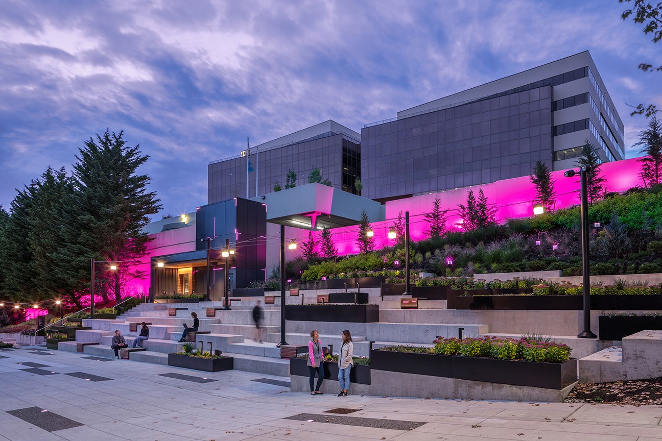 The outdoor amphitheater at dusk, aglow with bold magenta "digit" lighting and surrounded by planters and wooden benches arranged throughout the tiers.