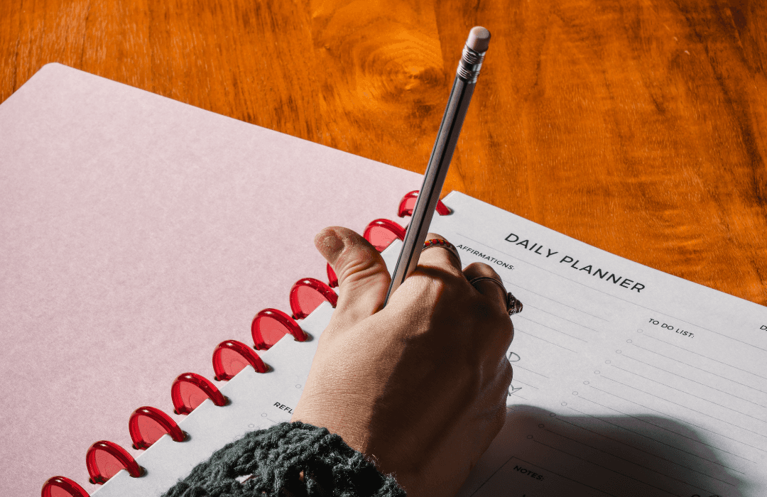 Someone holding a pencil writing in a daily planner. This planner has disks in the binding that make it so you can take the paper in and out without ripping any paper. The notebook is open on a wood table.