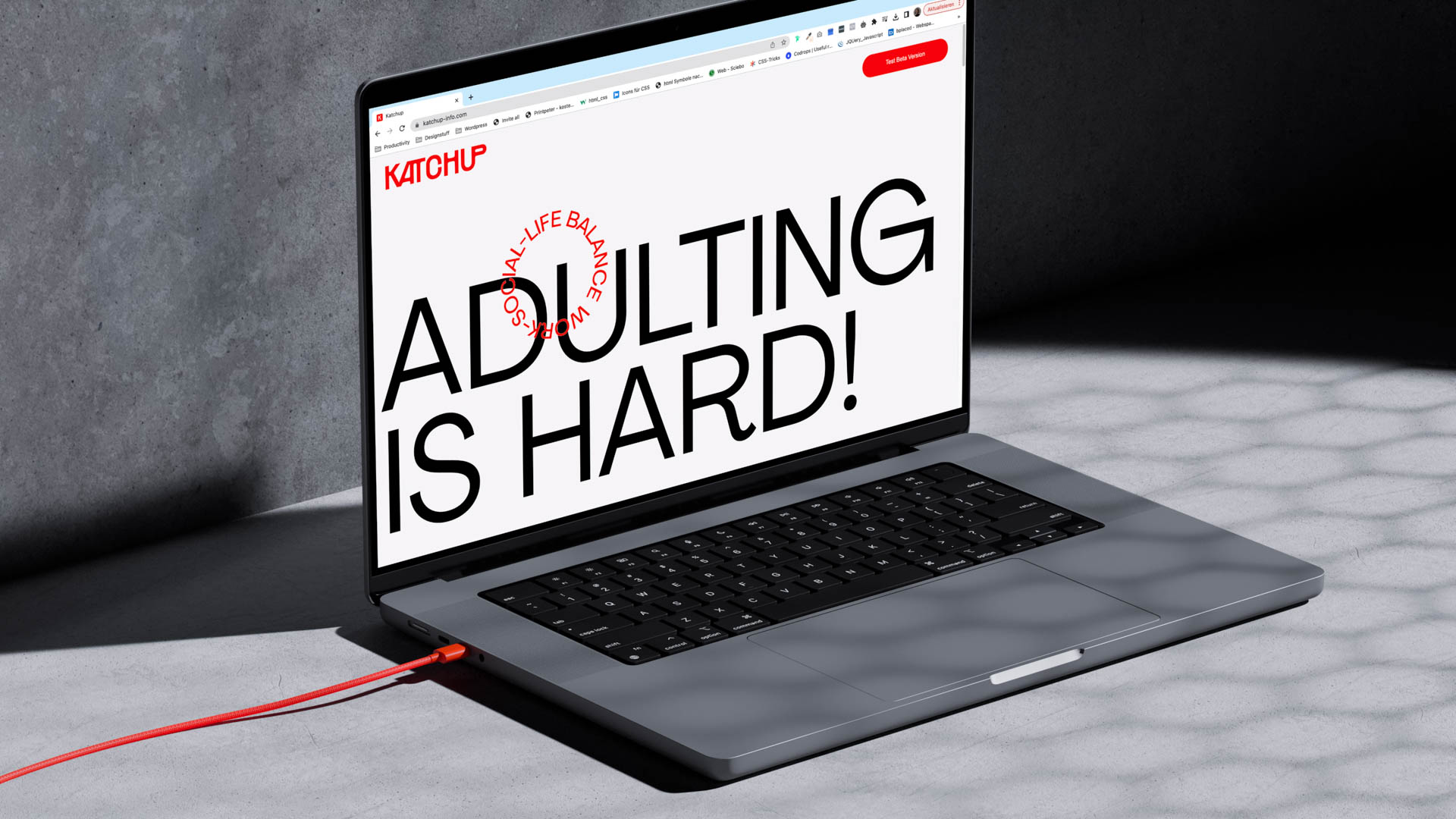A dark gray laptop computer sits on a light gray surface with a subtle hexagonal pattern. The screen displays a white background with the text "ADULTING IS HARD!" in bold, black, sans-serif font. A red circular logo reading “SOCIAL-LIFE-BALANCE” is visible in the upper left corner of the screen. A red cable extends from the side of the laptop, partially visible against the surface. The laptop is angled slightly to the right, and the lighting casts a shadow to the left of the device.