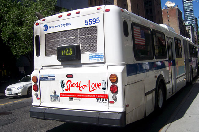 A white New York City bus, numbered 5595, is parked on a city street. The rear of the bus displays a digital sign reading "M23," a red "Speak of Love" sticker, and blue text including a phone number. A small American flag is visible on the side window. A white car is parked nearby, and buildings are visible in the background.