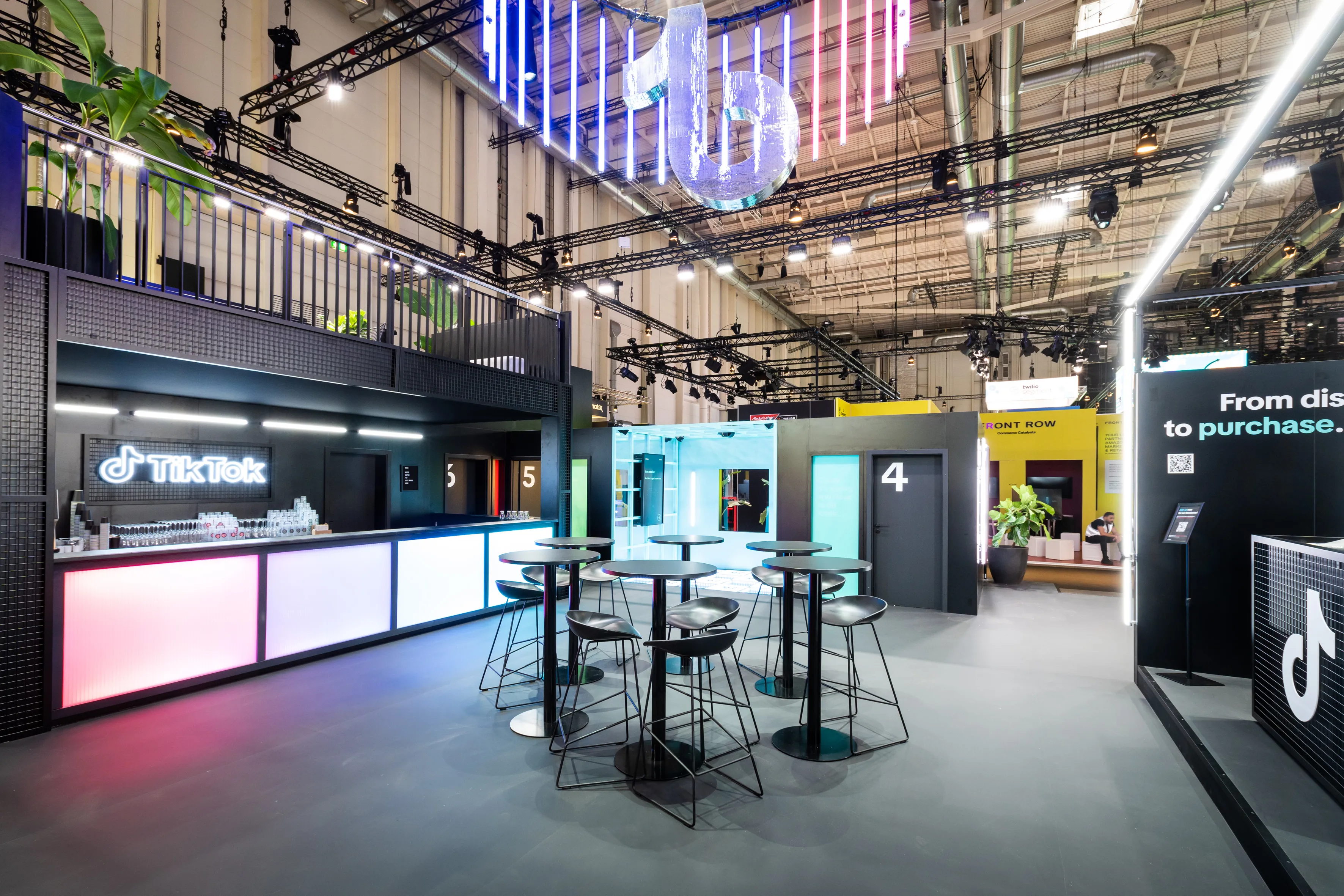 Bar with color-glowing counter and TikTok logo, high tables and numbered doors inside the OMR booth.
