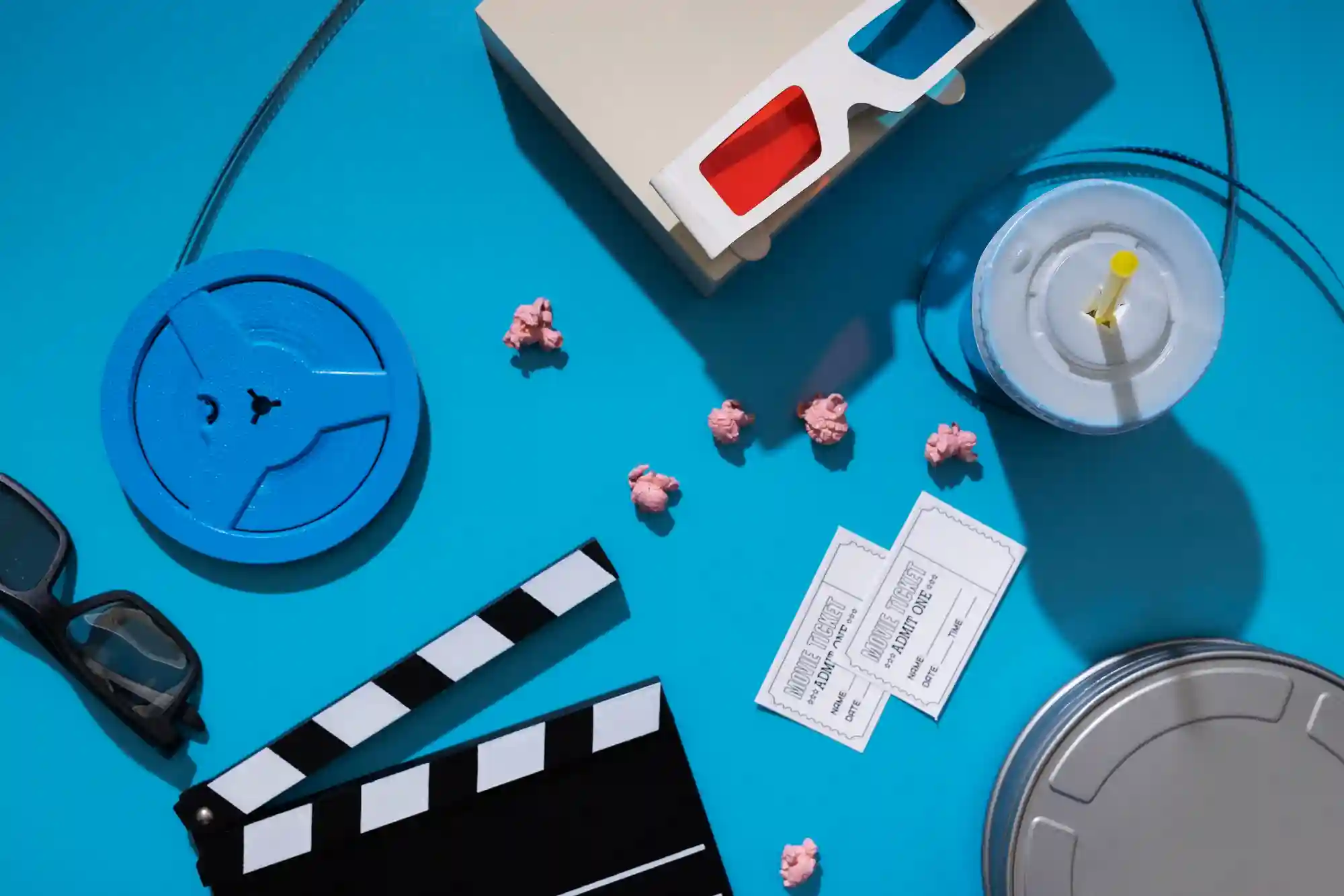 A flat lay of cinema essentials including a film reel, popcorn, 3D glasses, and tickets on a blue background.