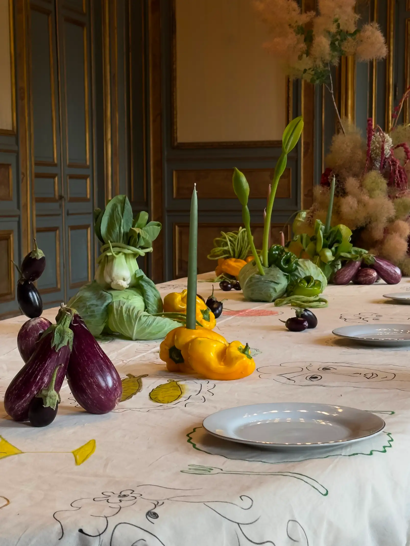 Mesa elegante decorada con un mantel ilustrado, adornada con berenjenas, pimientos amarillos, repollos y un pak choi atado como figura, junto a velas verdes y platos blancos, en un salón clásico con paredes de madera y detalles dorados