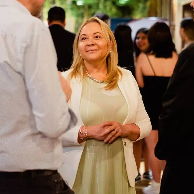 A woman in a white jacket and light green dress smiles while engaging in conversation at a social gathering. The atmosphere is warm and friendly.