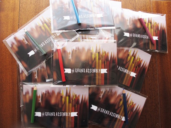 Several clear plastic pencil cases are arranged on a wooden surface. Each case contains a collection of colored pencils and has a white label with the text "THE GRAND ASSEMBLY" printed on it. The pencils are arranged in various colors and orientations within the cases.