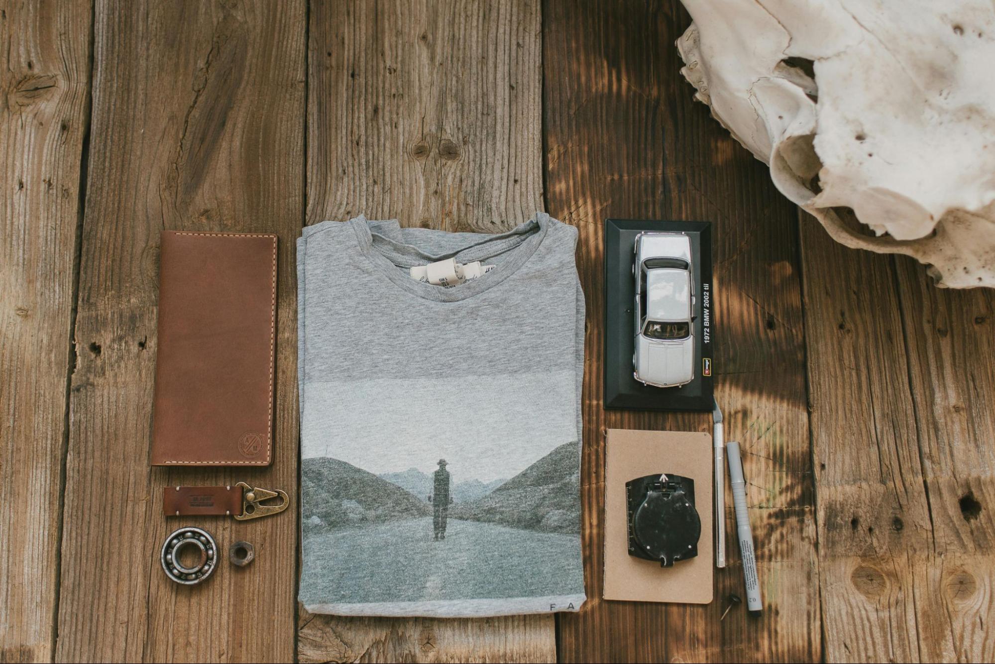 A wooden tabletop upon which various objects are neatly arranged. Starting from the left, there's a brown leather wallet with a metal clasp, a small white bear-shaped keychain, a round metal bearing, a pair of black pens, a notebook, a compass, a folded gray t-shirt with an image of a person standing on a road amidst mountains, and a miniature model of a silver car. To the right of the t-shirt, there's a white, weathered skull.