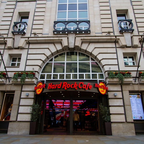 Front entrance of a Hard Rock Cafe with its lit red sign, and the interior visible through open doors under a large arched window.