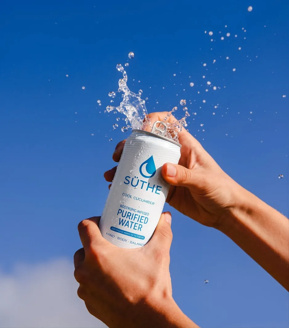 Hands open a SÜTHE purified water can under a bright blue sky, with water splashing up.