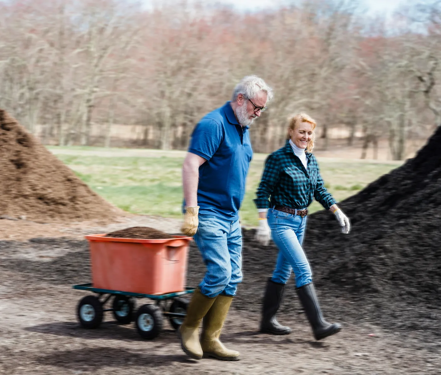 Two older people in gumboots transporting dirt from piles in an orange wagon.