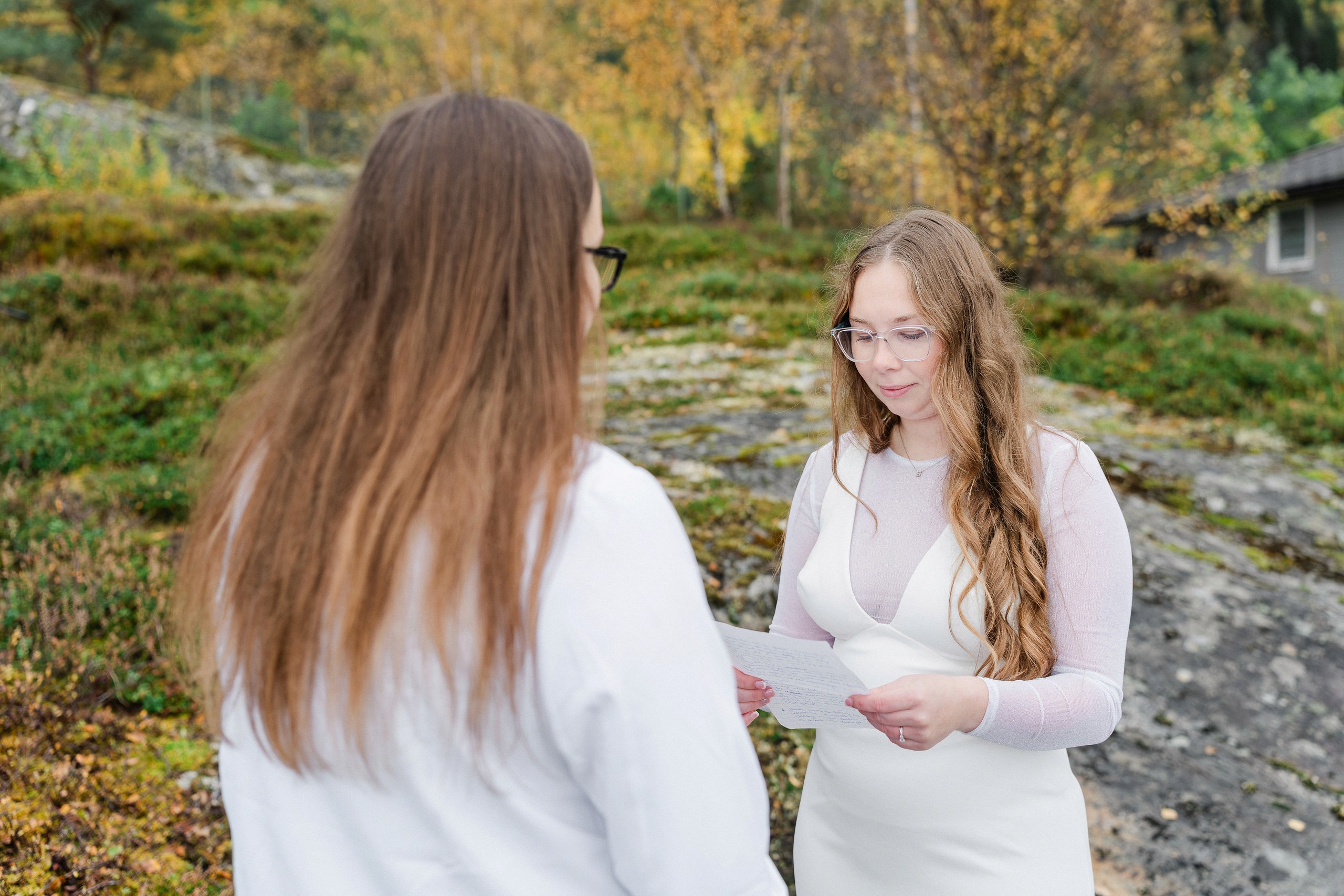 One partner reads from a sheet while the other stands in the foreground, surrounded by rocky ground, greenery and a small building.