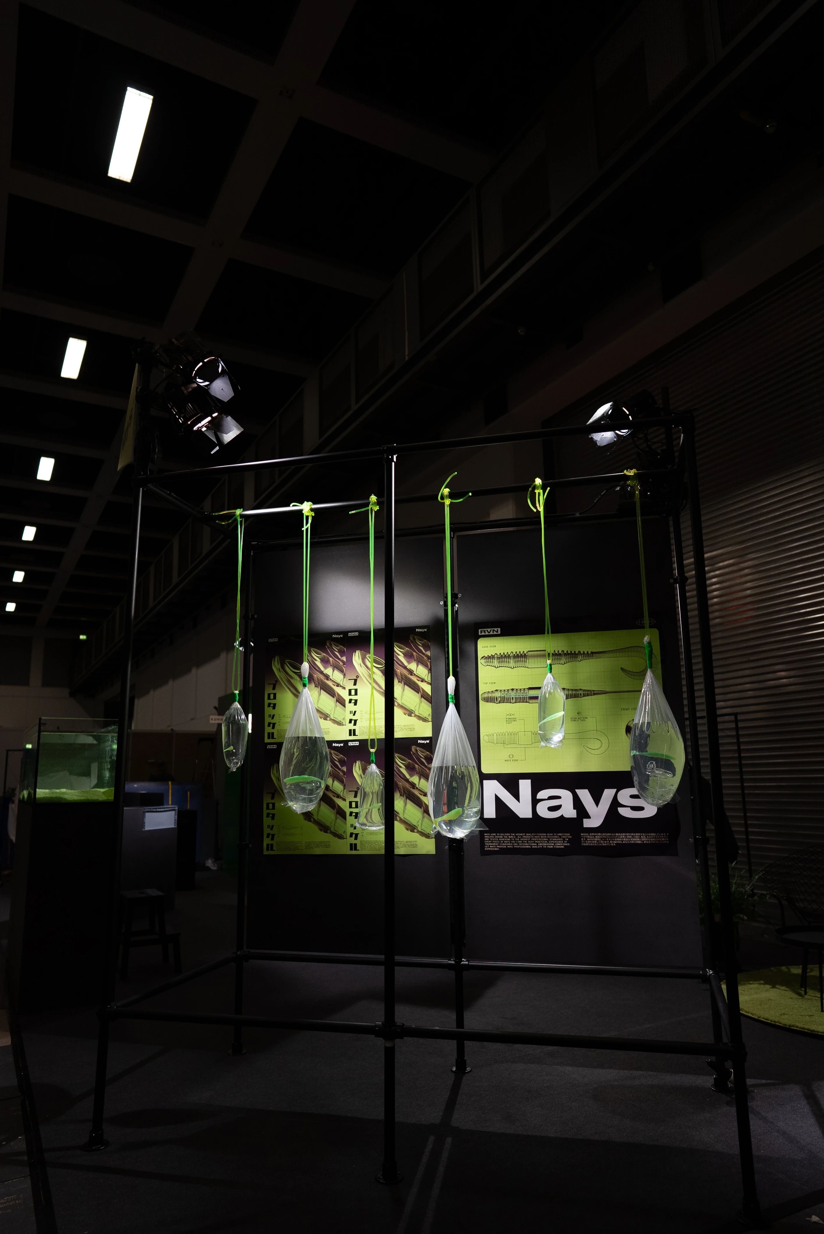 Installation of water-filled bags with lures dangling from a black frame in front of the Nays poster wall.