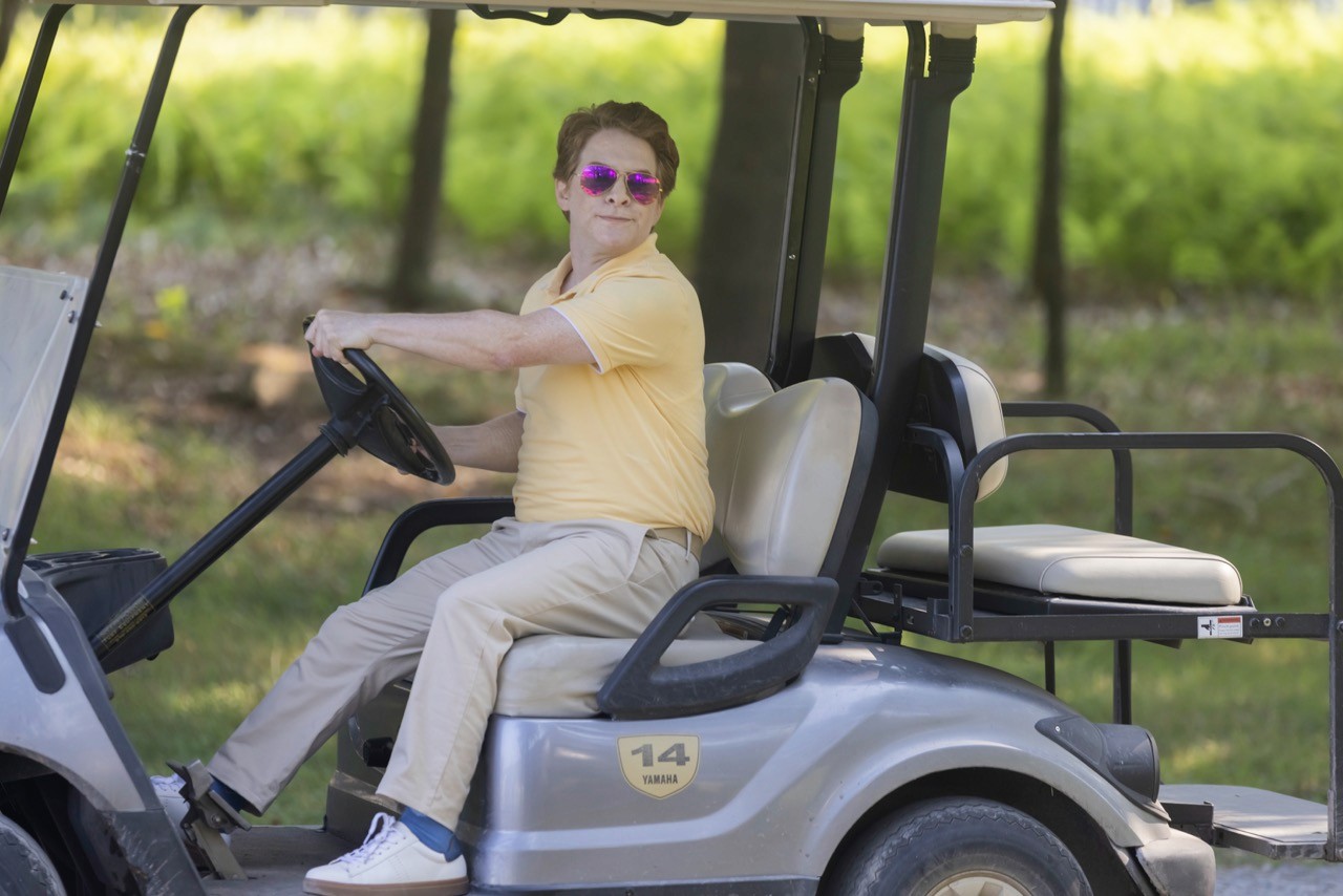 Still from The Floaters: A man in a yellow polo shirt and purple-tinted sunglasses drives a golf cart with a smug expression.