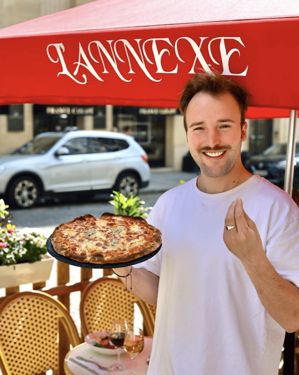 Person with a mustache wearing a black t-shirt carrying a tall stack of pizza boxes inside a dimly lit bar or restaurant.