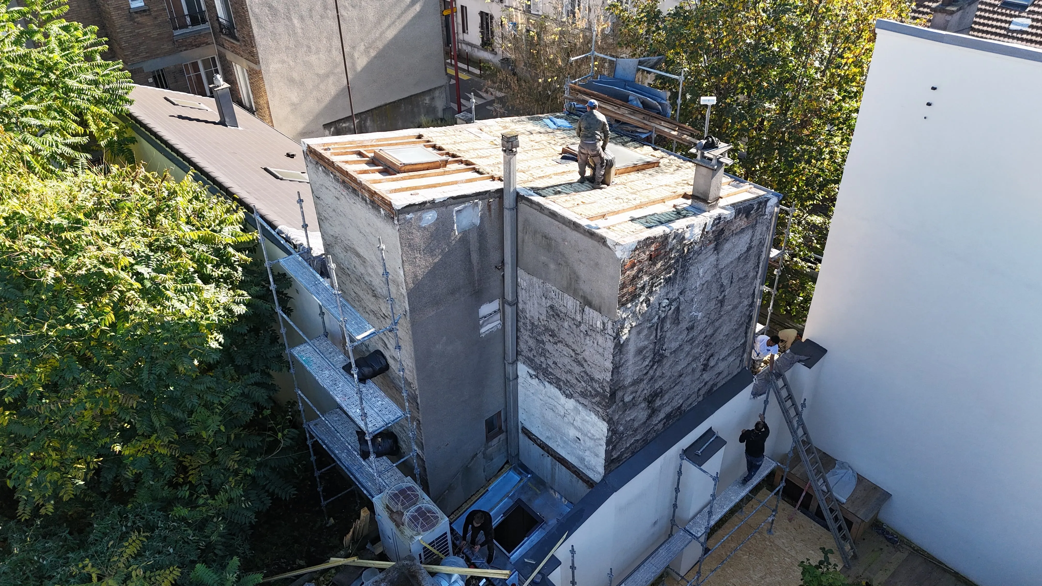 Vue plongeante sur une toiture partiellement découverte, avec une structure en bois apparente, deux ouvertures vitrées et un échafaudage
