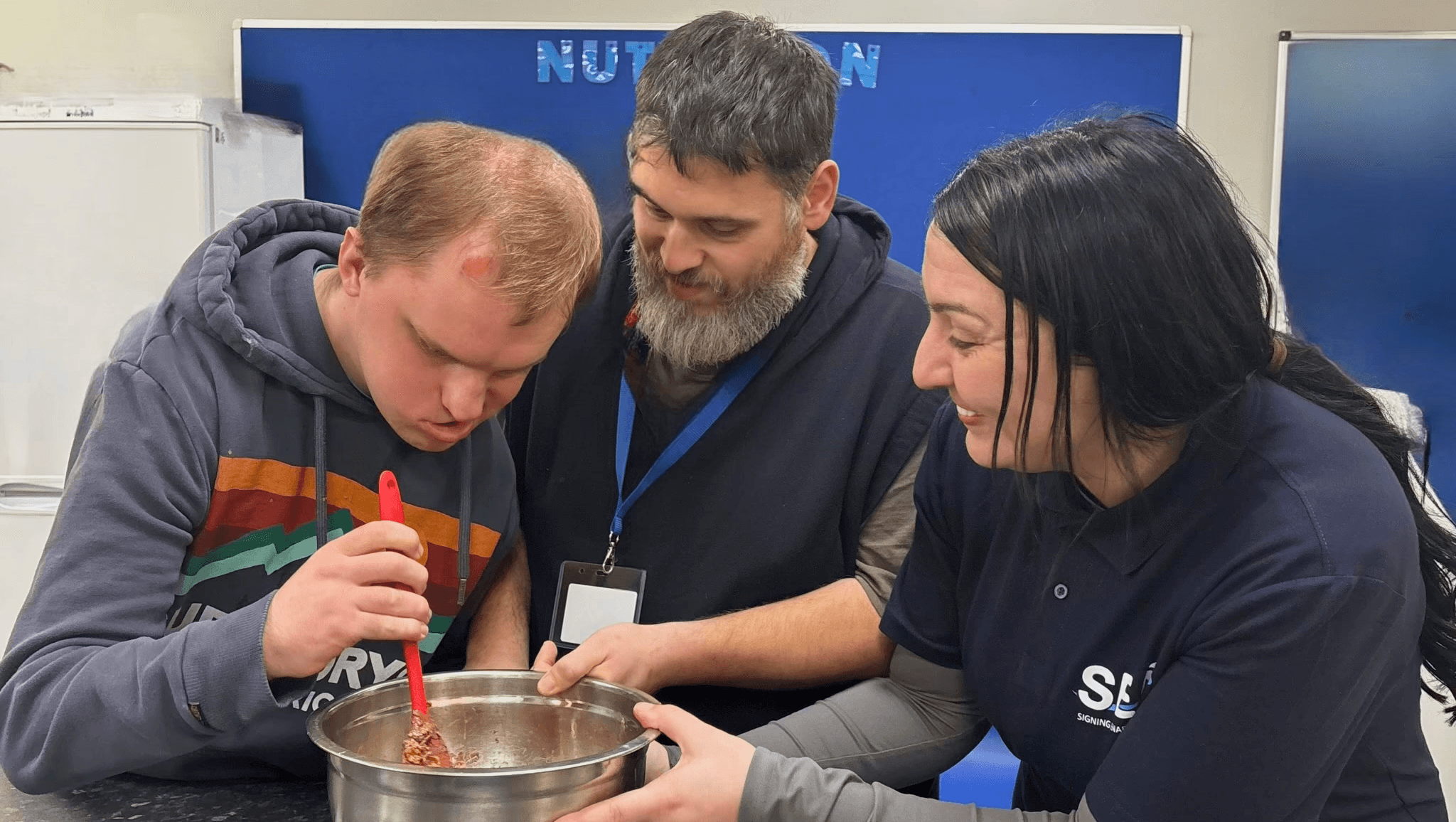 A support worker assists a young man with a cooking activity, mixing ingredients in a bowl together at a table