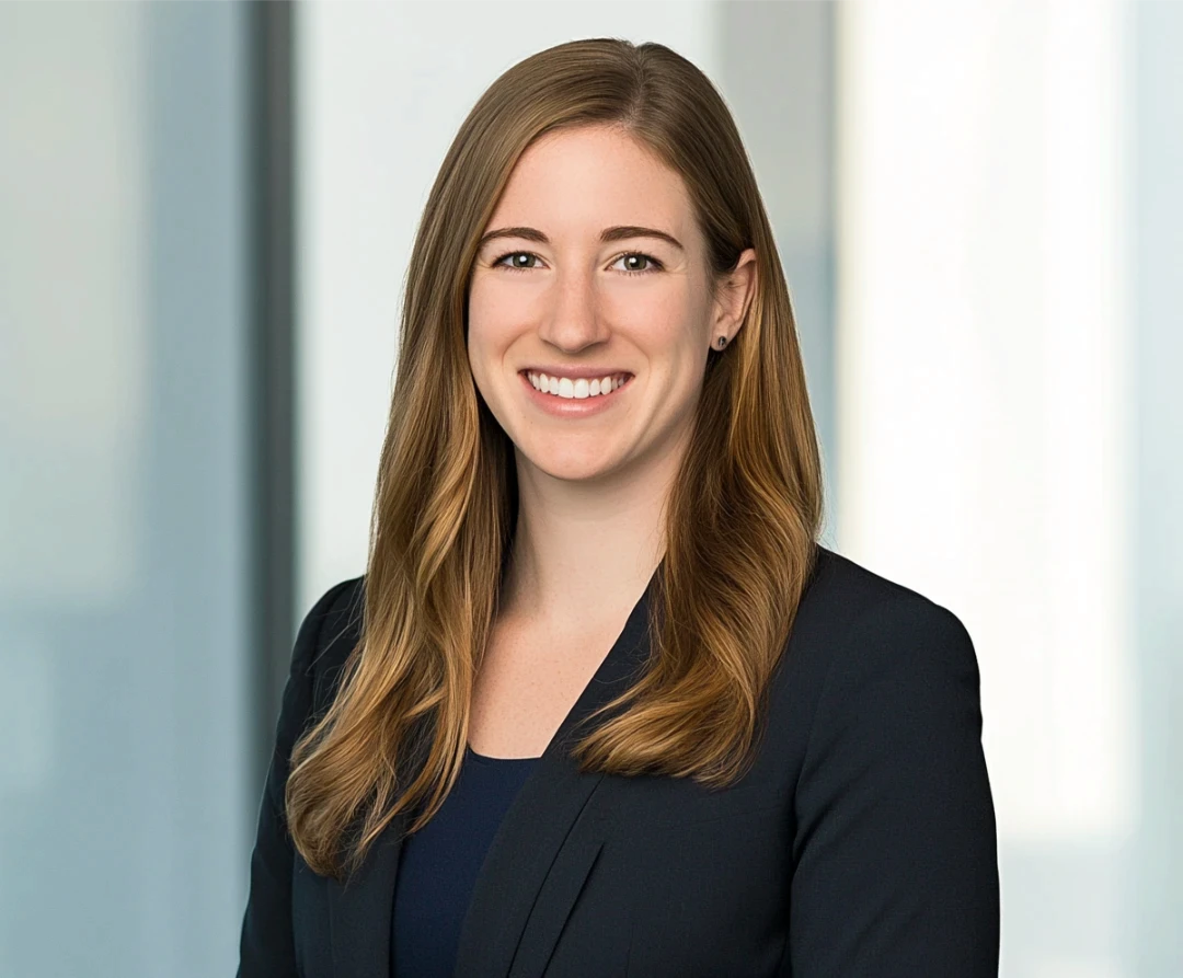 Smiling woman with long brown hair wearing a dark blazer stands in a bright, modern office setting. She exudes confidence and professionalism.