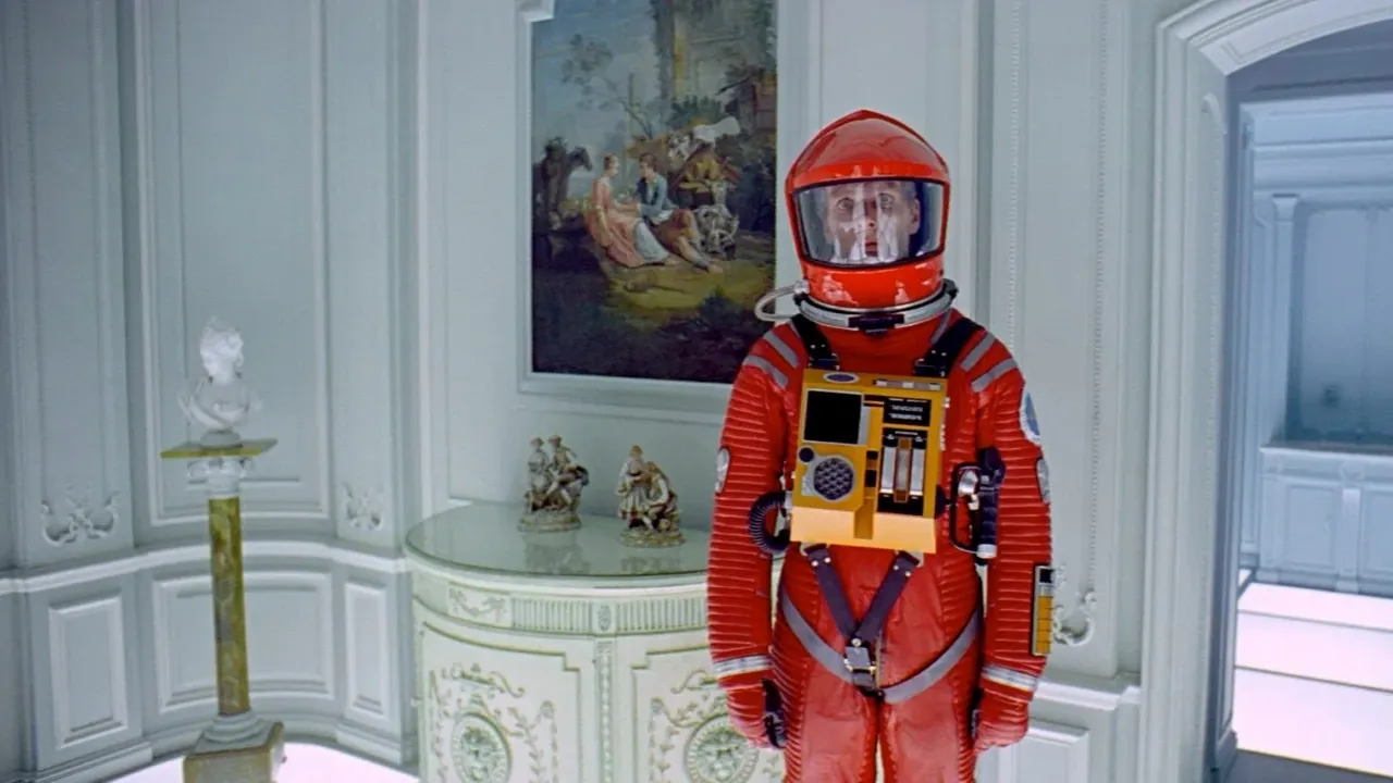 A person in a red space suit stands in an elegantly decorated room with white walls and ornate furniture, featuring a classical painting and decorative figurines.