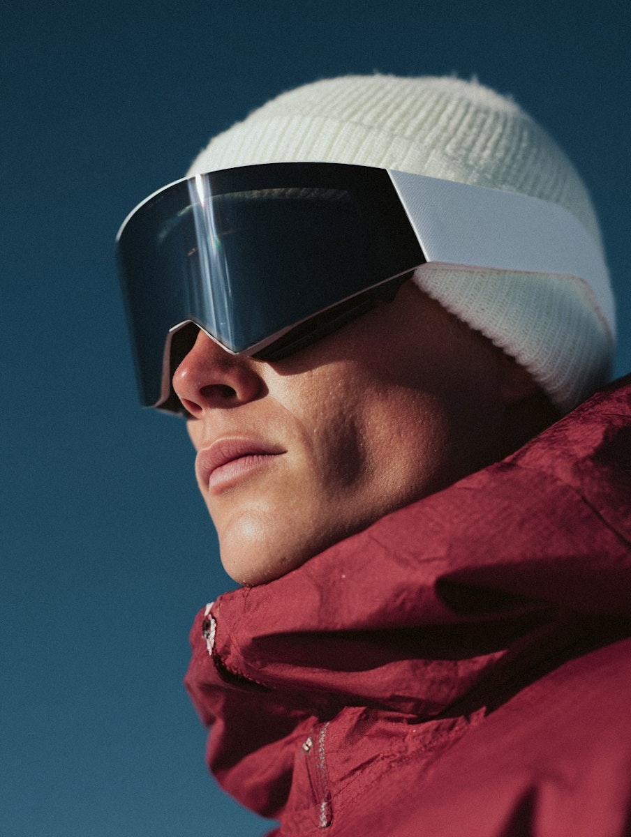 Side-profile portrait of a person wearing a white beanie, oversized black ski goggles, and a red insulated jacket against a clear blue background.