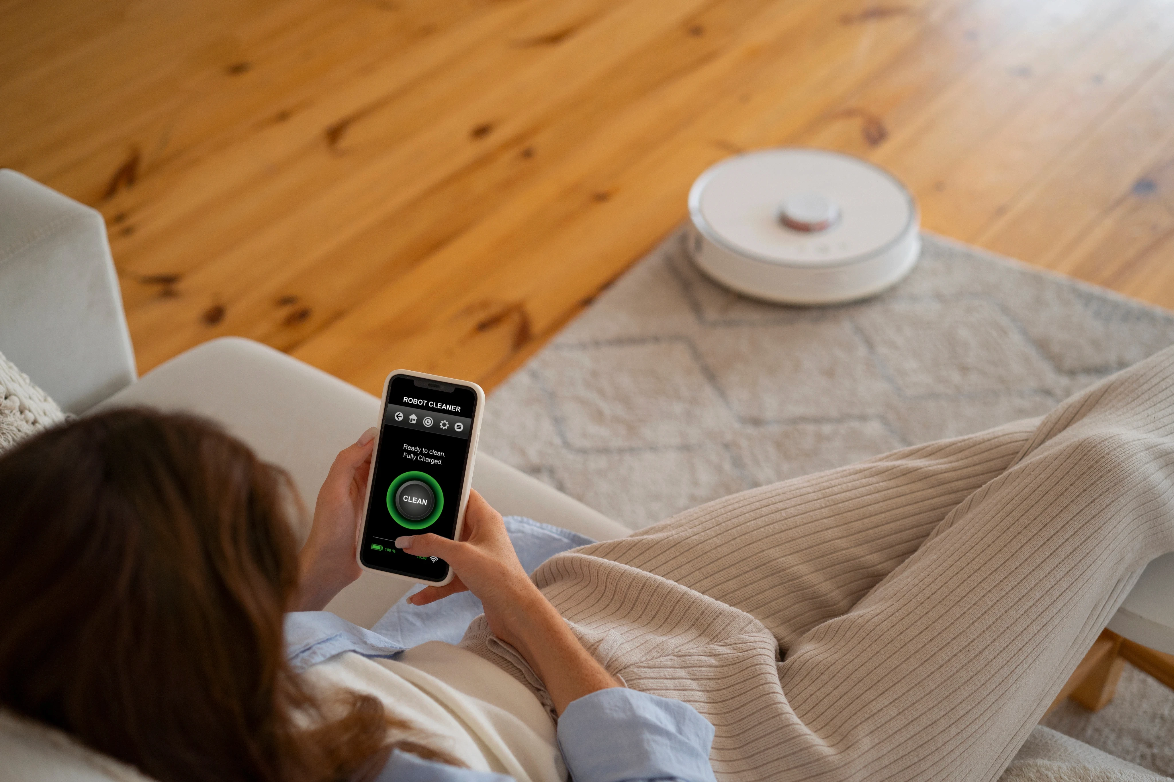 Person using a smartphone app to coordinate smart devices and a robot vacuum on a rug.
