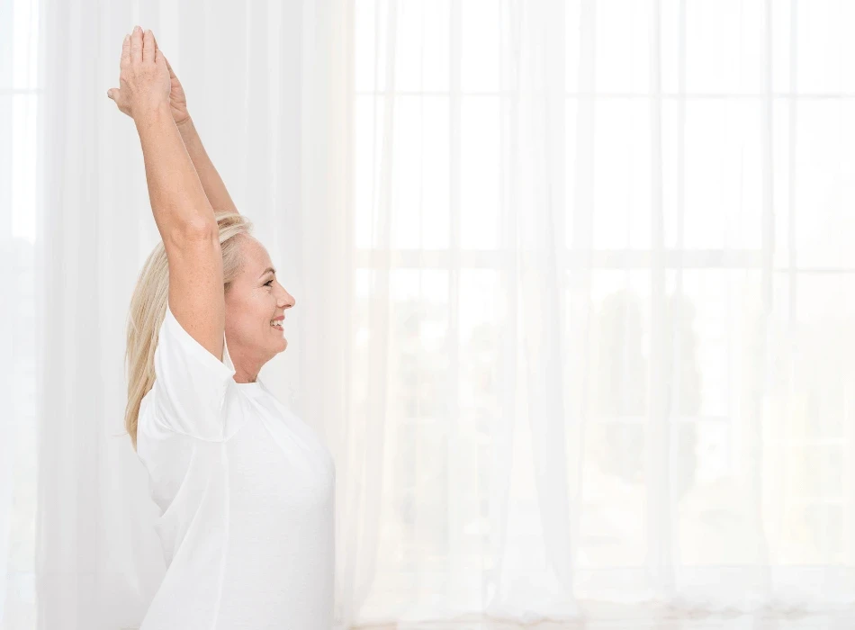 Woman in a white shirt doing an overhead stretch by a bright window, concept for pelvic pain and period management.